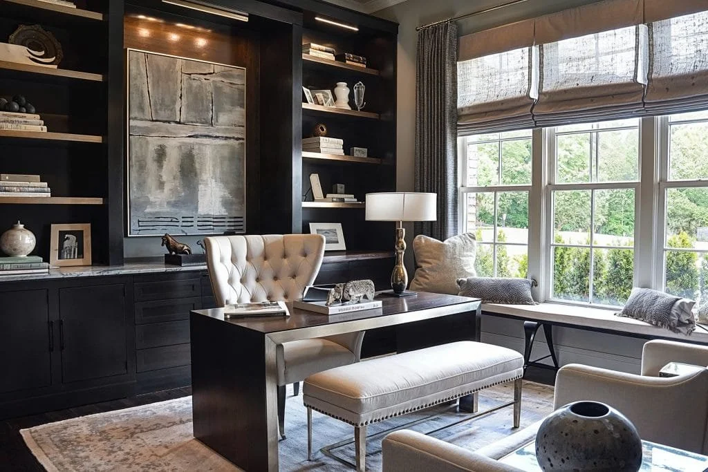 A well-decorated home office or reading nook with a dark wooden built-in bookshelf filled with books and decorative items, a window seat with throw pillows, a large window with Roman shades allowing natural light, a desk with a white upholstered chai