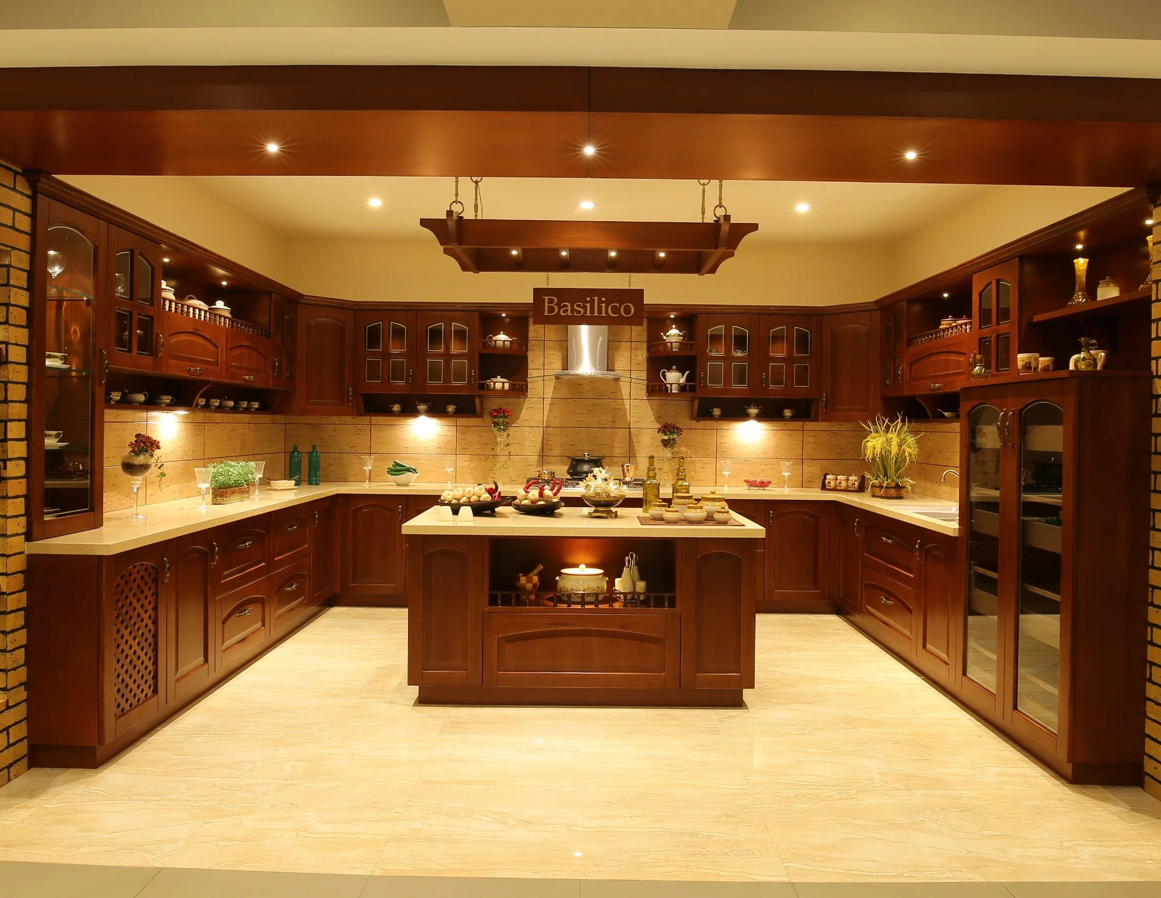 A warm, well-lit kitchen with wooden cabinets, a central island, and a sign that says 'Basilico.' The kitchen features various dishes, plants, and decorative items on the countertops and shelves.