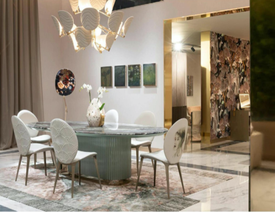 Modern dining room with a large round table, white upholstered chairs, a floral chandelier, art on the walls, and a marble floor.