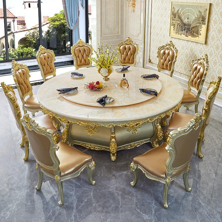 Elegant dining room with an ornate round marble table surrounded by nine gold-trimmed upholstered chairs. The table is set with some plates, a vase of yellow flowers, a bowl of fruit, and a small bottle of perfume. Large windows with blue curtains re
