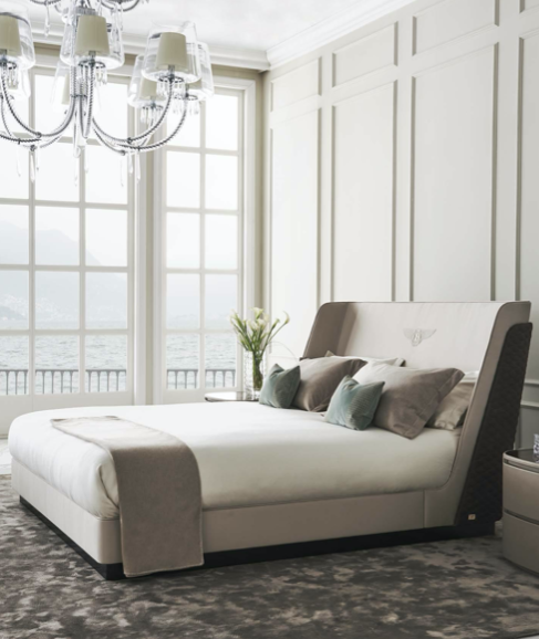 Elegant bedroom with a large bed, decorative pillows, bedside table with a vase of flowers, large windows with ocean view, and a chandelier.