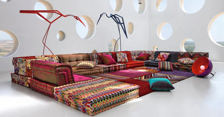 A large, colorful sectional sofa with patterned cushions arranged in a modern room with circular cutouts on the white wall and artistic floor lamps overhead.