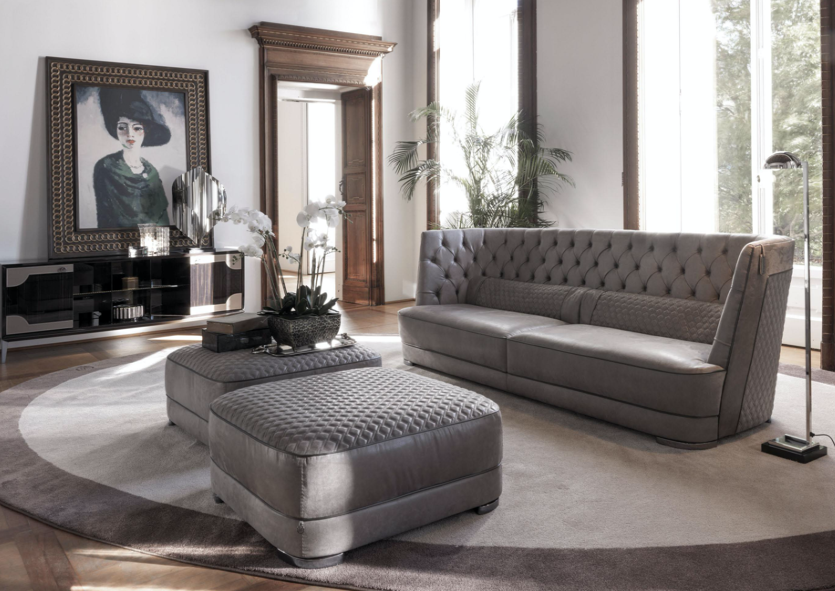 A modern living room with a gray tufted sofa, upholstered ottomans, a circular area rug, a large painting of a woman in a hat, a console table with decorative items, a standing floor lamp, and large windows letting in natural light.