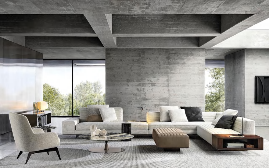 Modern living room with concrete walls, large window showing trees outside, white sectional sofa with black and white accent pillows, beige armchair, round coffee table with decorative objects, brown ottoman, and flat-screen TV on the wall.