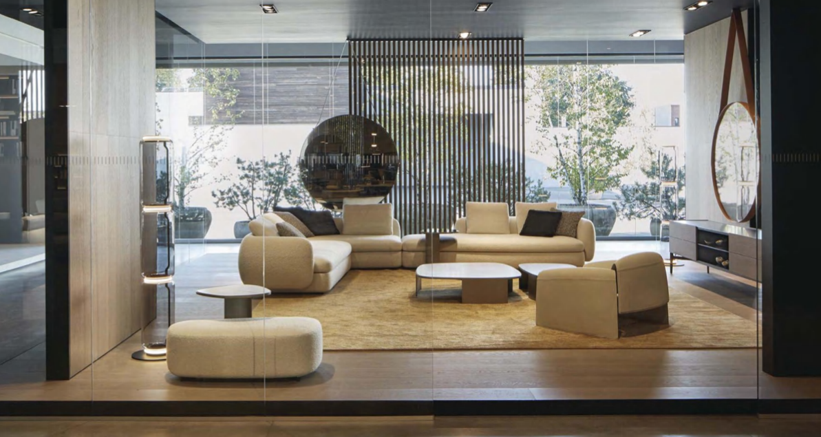 Modern hotel lobby with cream-colored sofas, a round hanging mirror, a wooden slatted divider, and large glass windows with trees outside.
