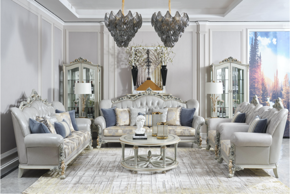 Elegant living room with white tufted sofas, decorative pillows, glass-top coffee table, ornate display cabinets, and a large window with an outdoor landscape view. Two modern chandeliers hang from the ceiling, and there are vases with tall flowers a