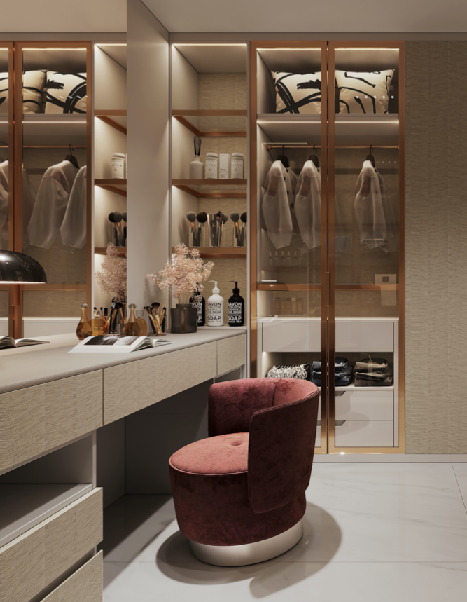Luxurious walk-in closet with beige cabinetry, glass-fronted wardrobe, and a velvet pink chair in front of a vanity with makeup brushes, skincare products, and decorative flowers.