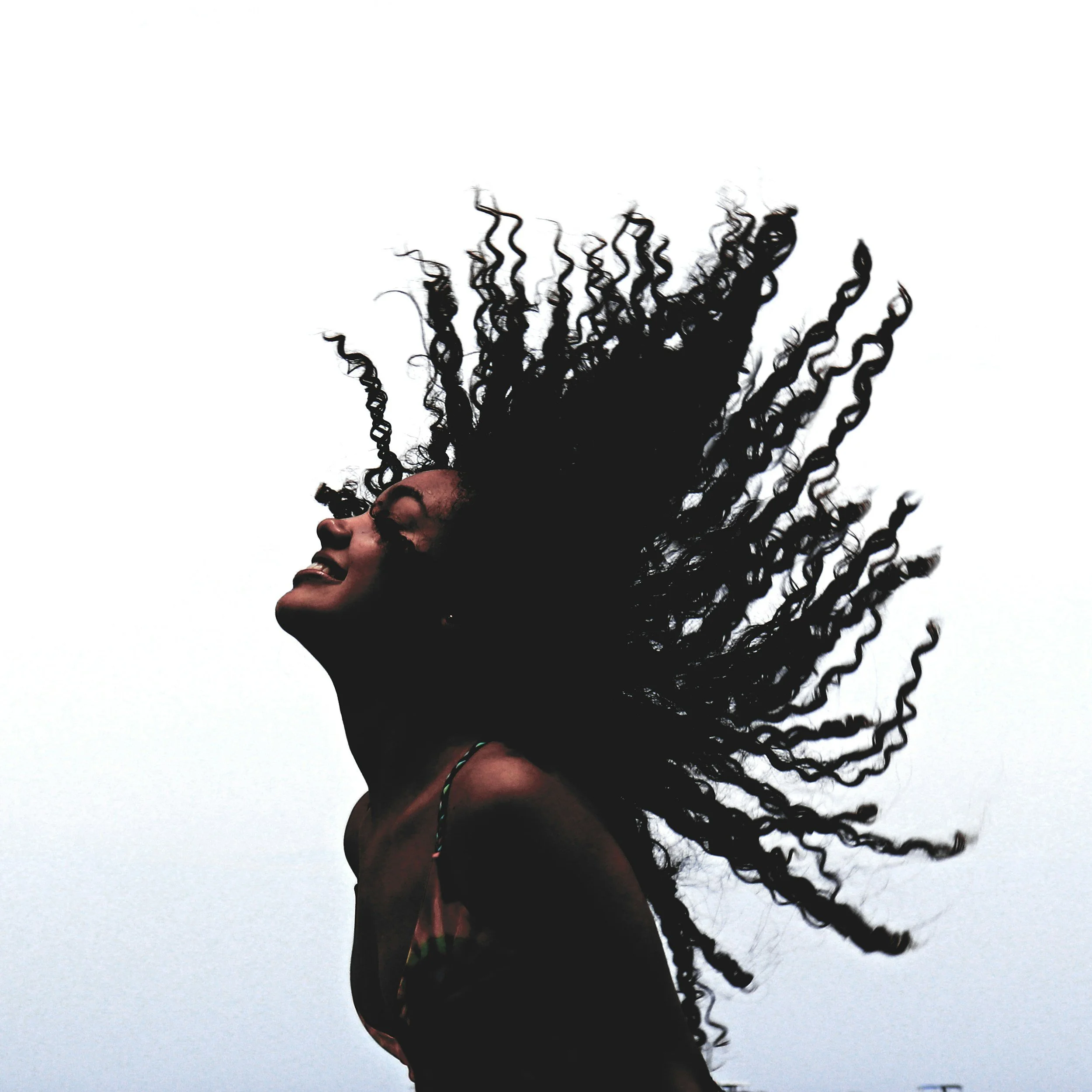 A woman with curly hair flipping back her hair with her head tilted upward and smiling against a cloudy sky.