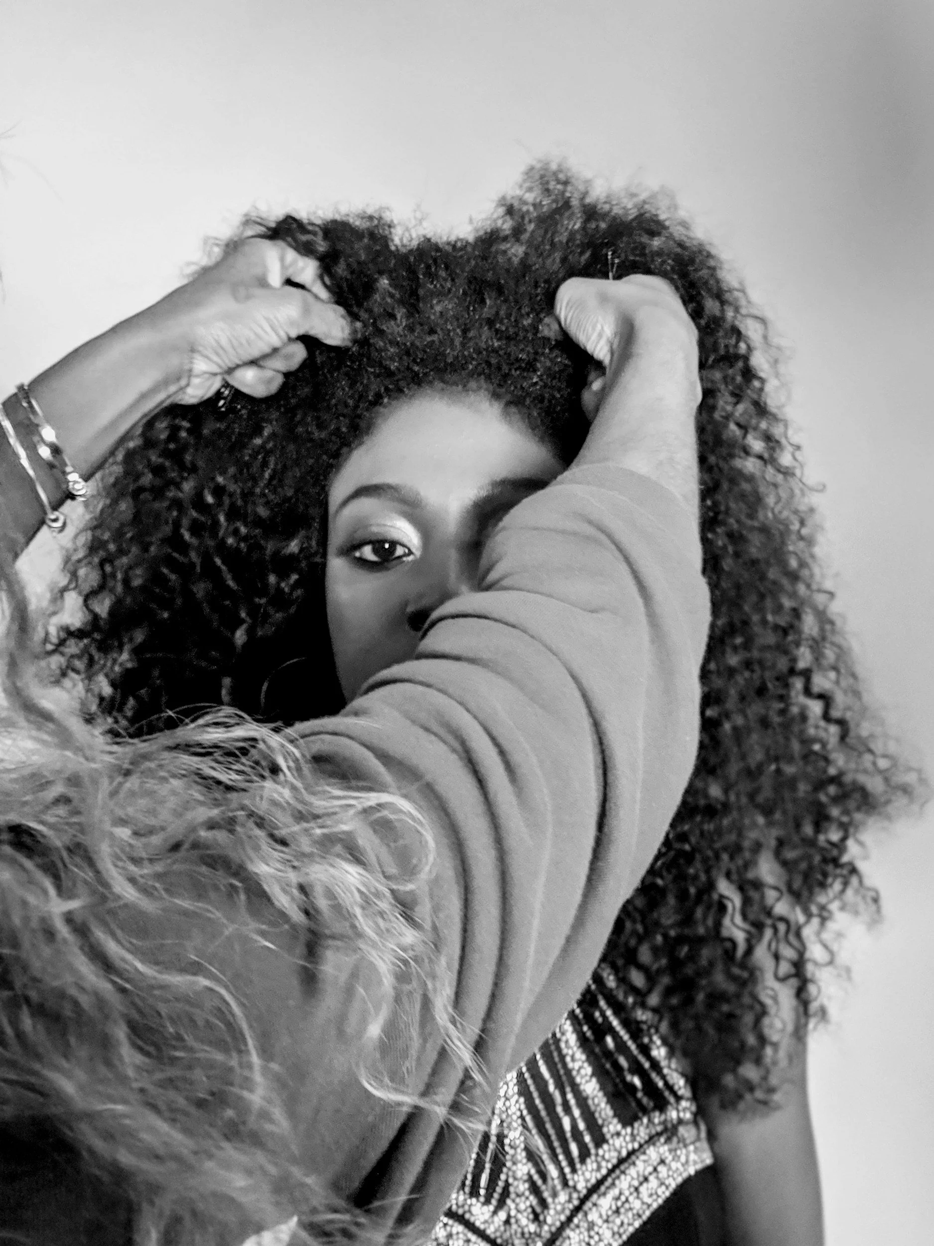 A woman with curly hair looks directly at the camera while someone adjusts or styles her hair.