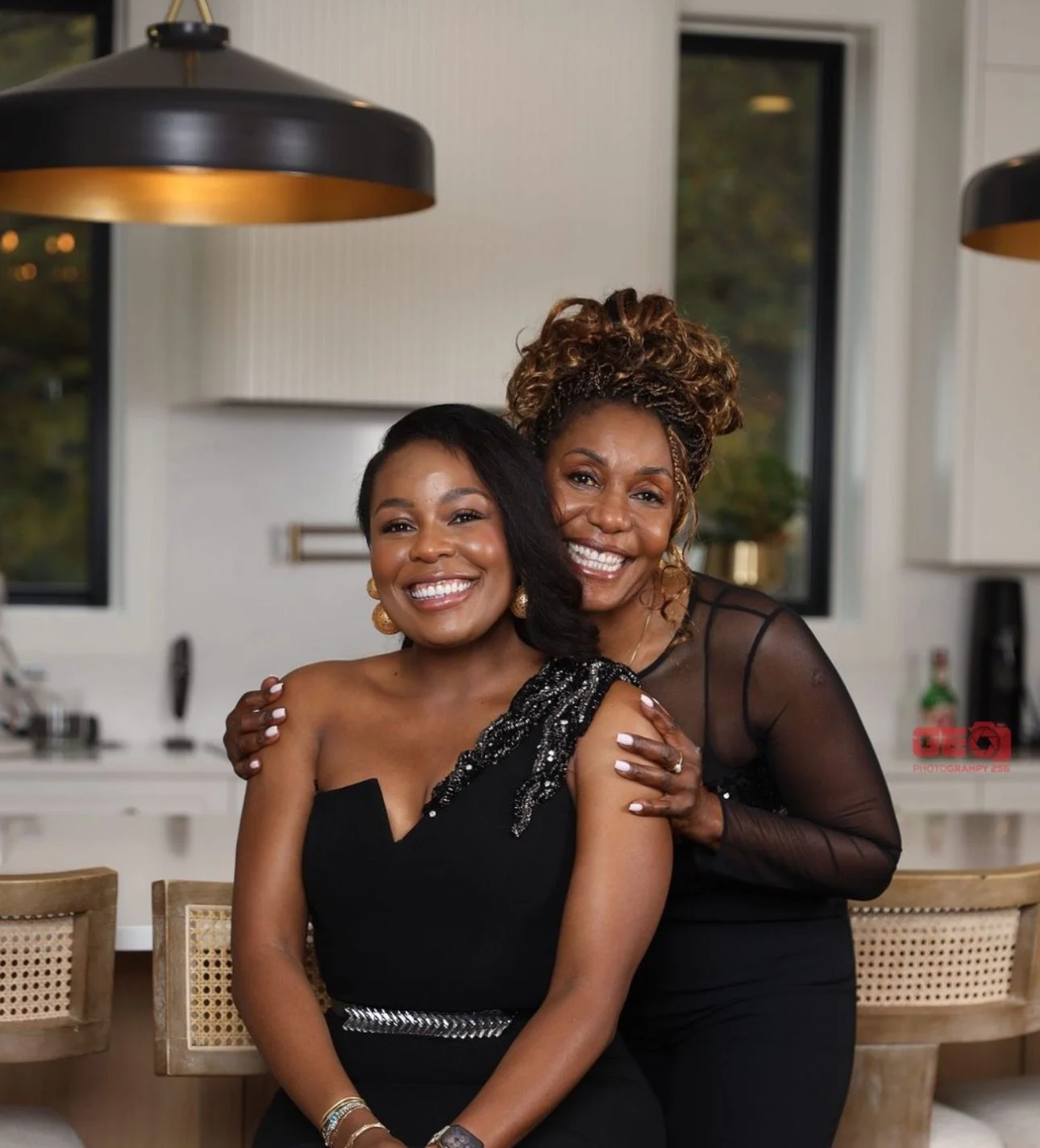 Two smiling women dressed in black, one with short hair and the other with curly hair and a sheer top, posing together in a well-lit modern kitchen.