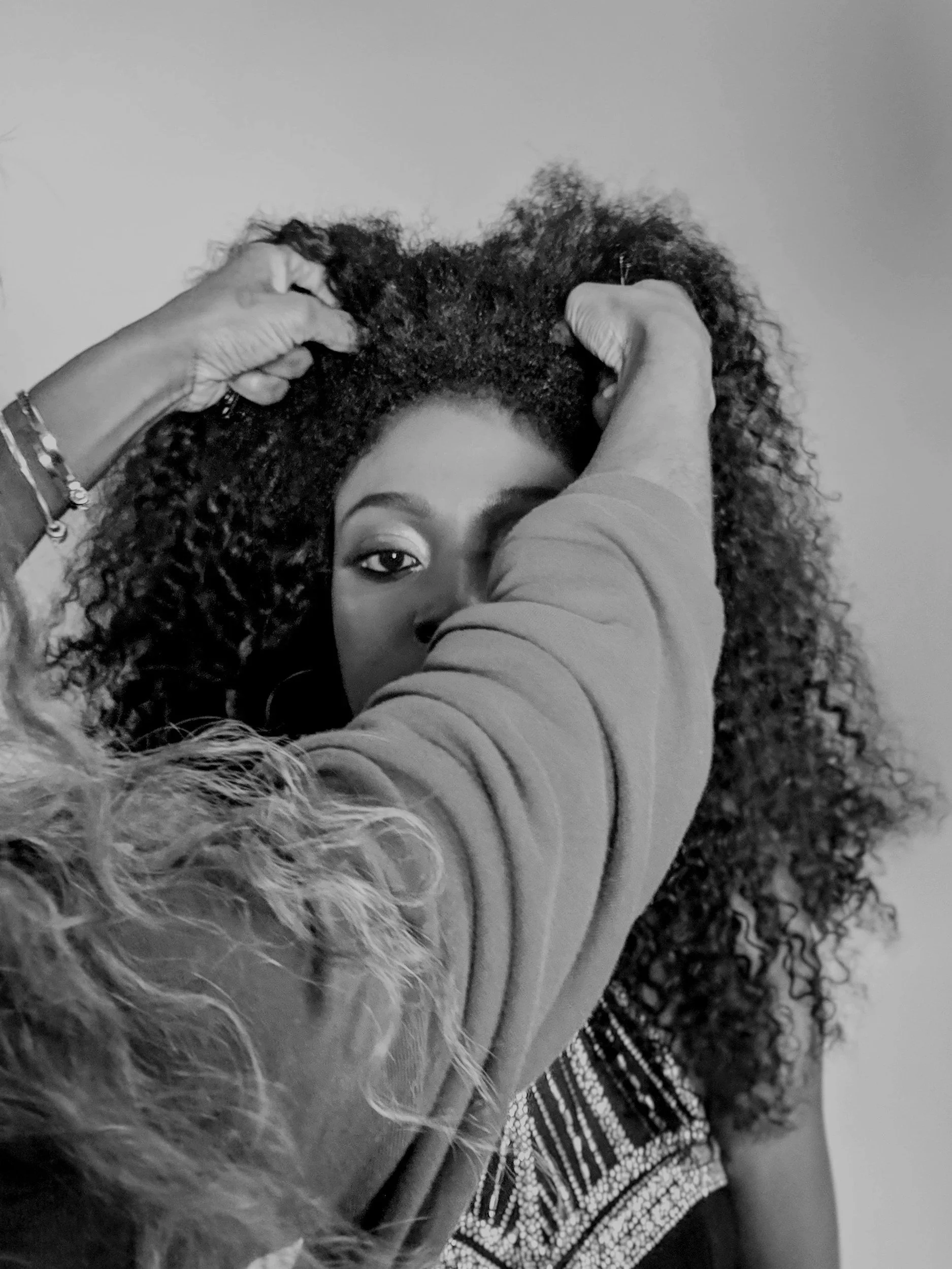 A person with curly hair holding the hair with both hands, wearing a long-sleeved shirt and bracelets. The person's face is partially visible, with one eye and part of the forehead showing. The background is plain and neutral.