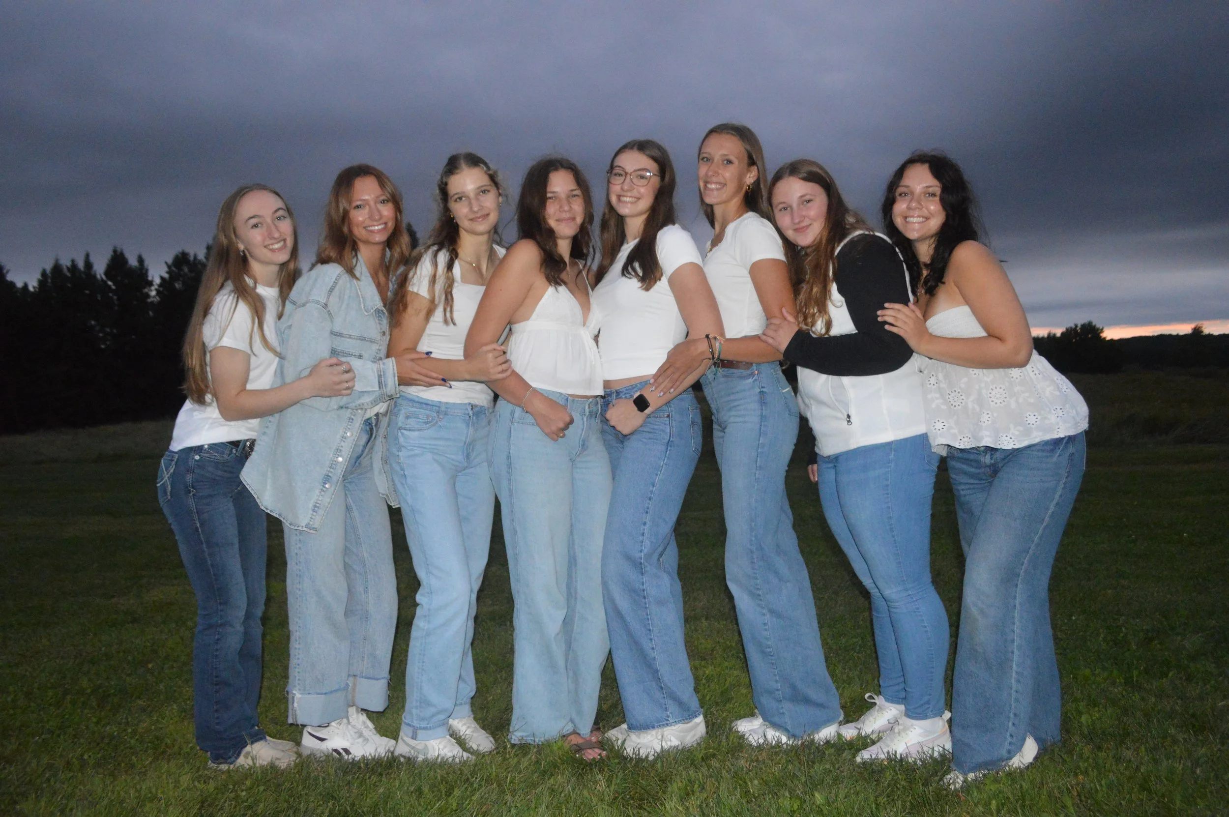 A group of nine young women standing outdoors on grass in front of a dark, cloudy sky at dusk. They are smiling and dressed in casual white and denim clothing, with some wearing sneakers.