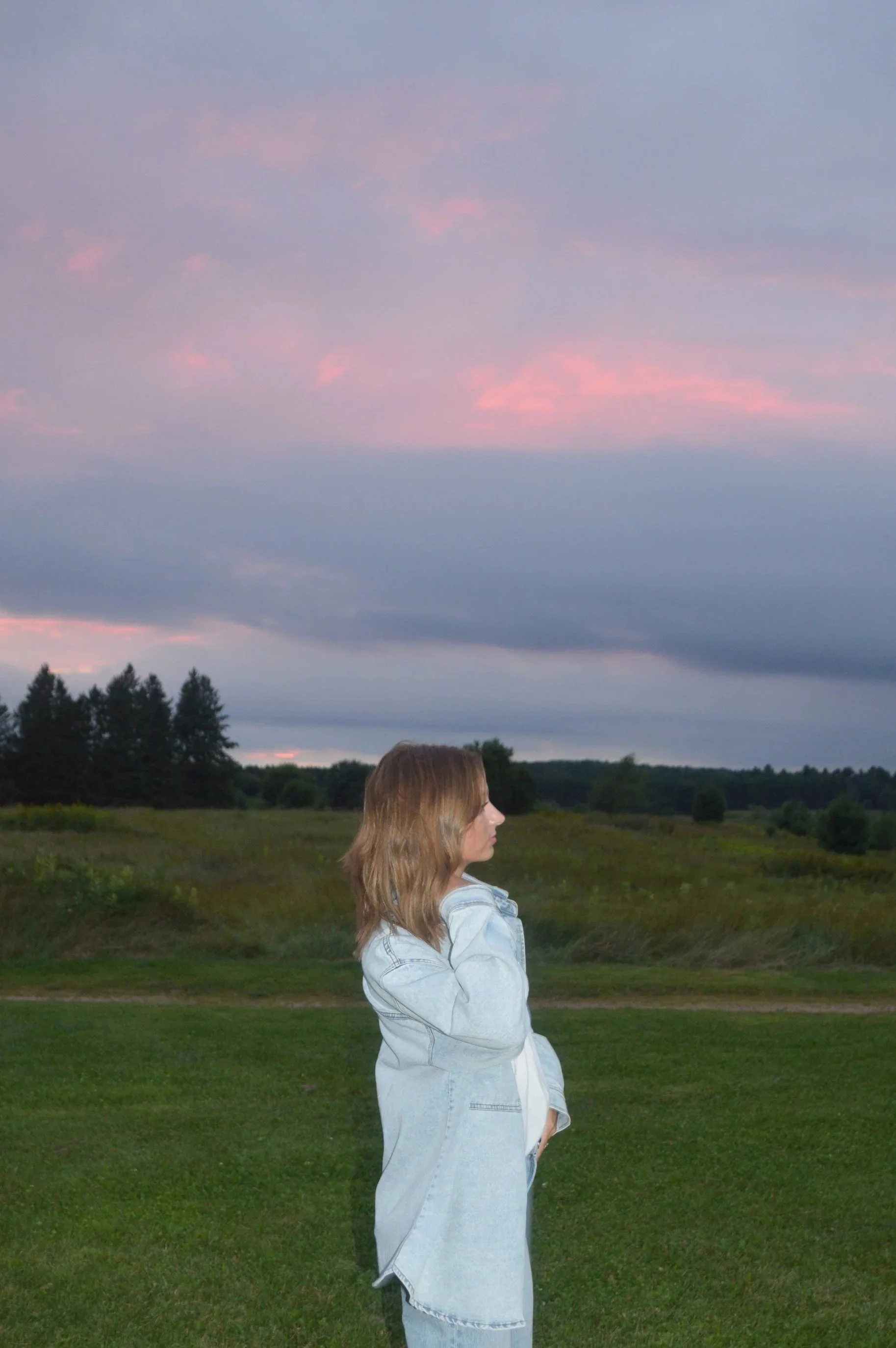 A young woman with blonde hair wearing a light blue denim jacket standing outdoors on grass, with a landscape of trees and hills in the background, under a partly cloudy sky during sunset.