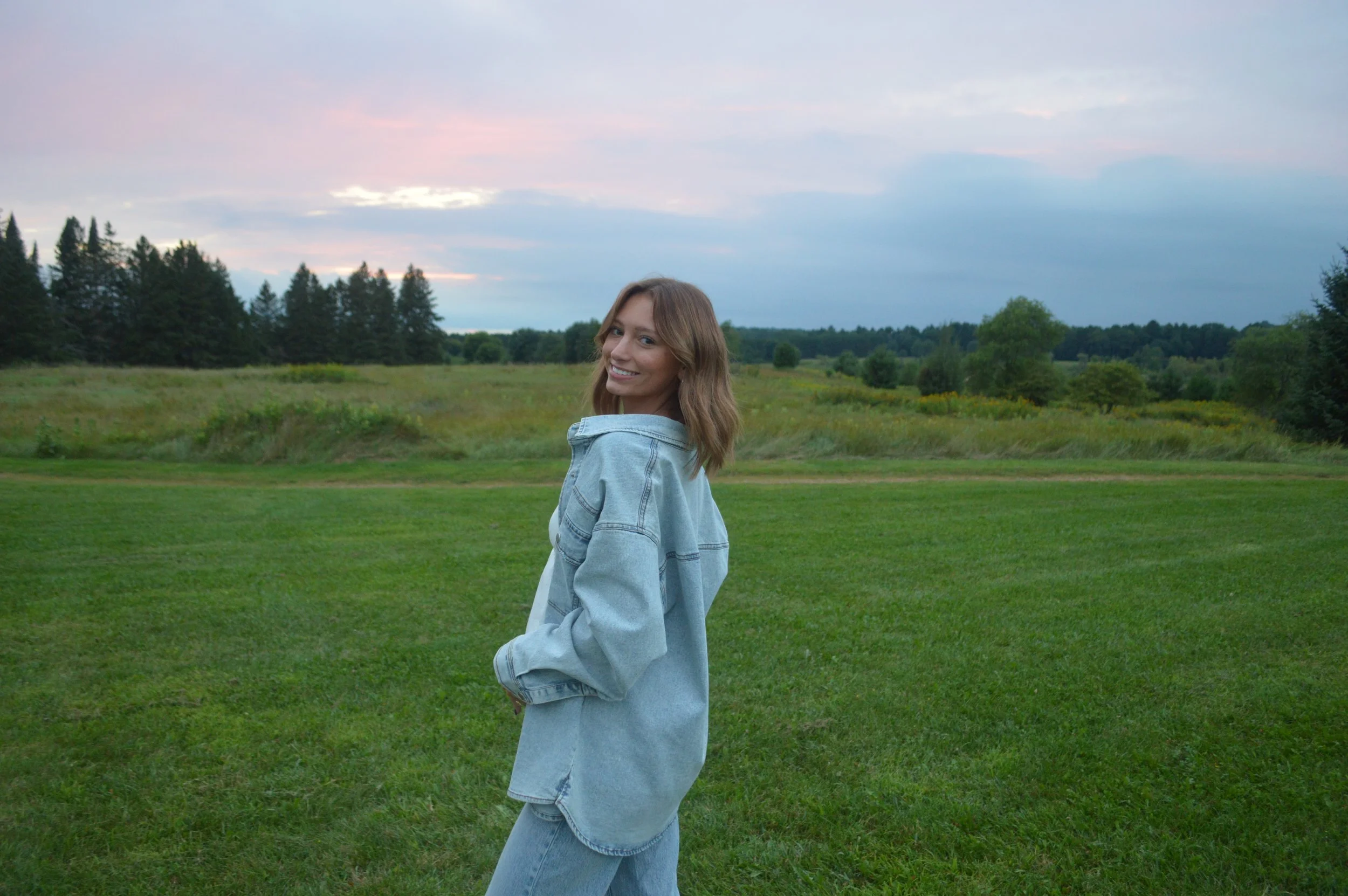 A young woman with shoulder-length brown hair smiling, wearing a light blue oversized denim jacket and matching jeans, standing in a lush green field with trees and a sunset sky in the background.