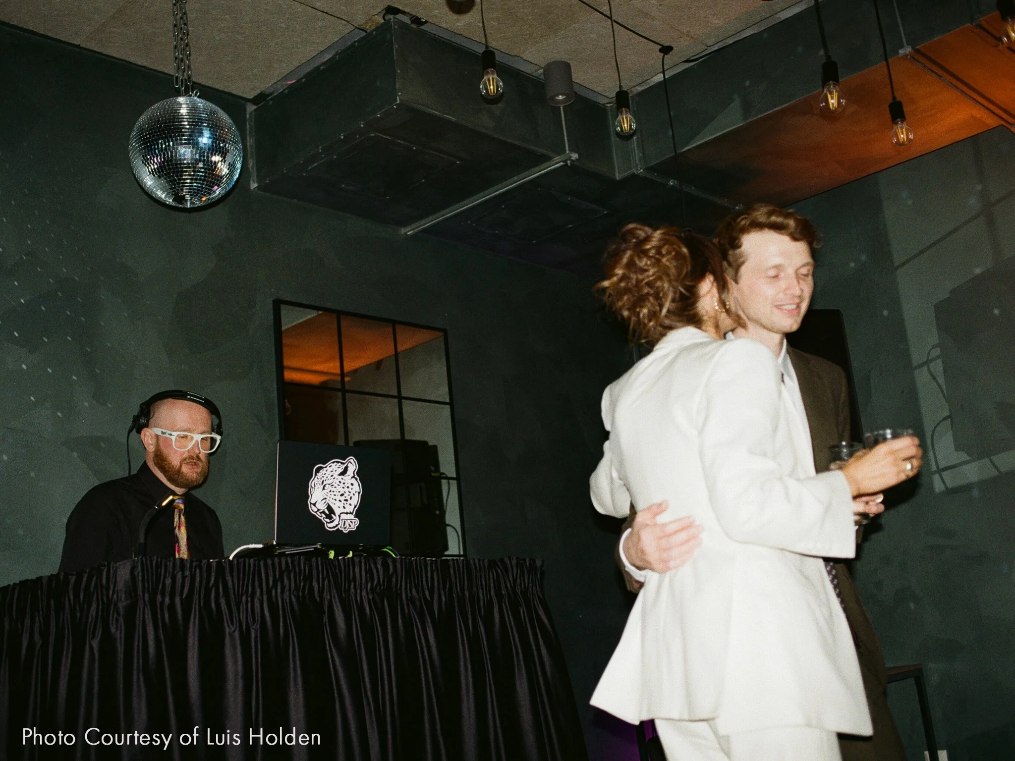 A couple dancing closely at a party with a DJ playing in the background. The woman has curly hair and is wearing a white blazer, while the man has short hair and is dressed in a suit. The DJ is wearing glasses and headphones, standing near a black table with a laptop that has a jaguar logo.