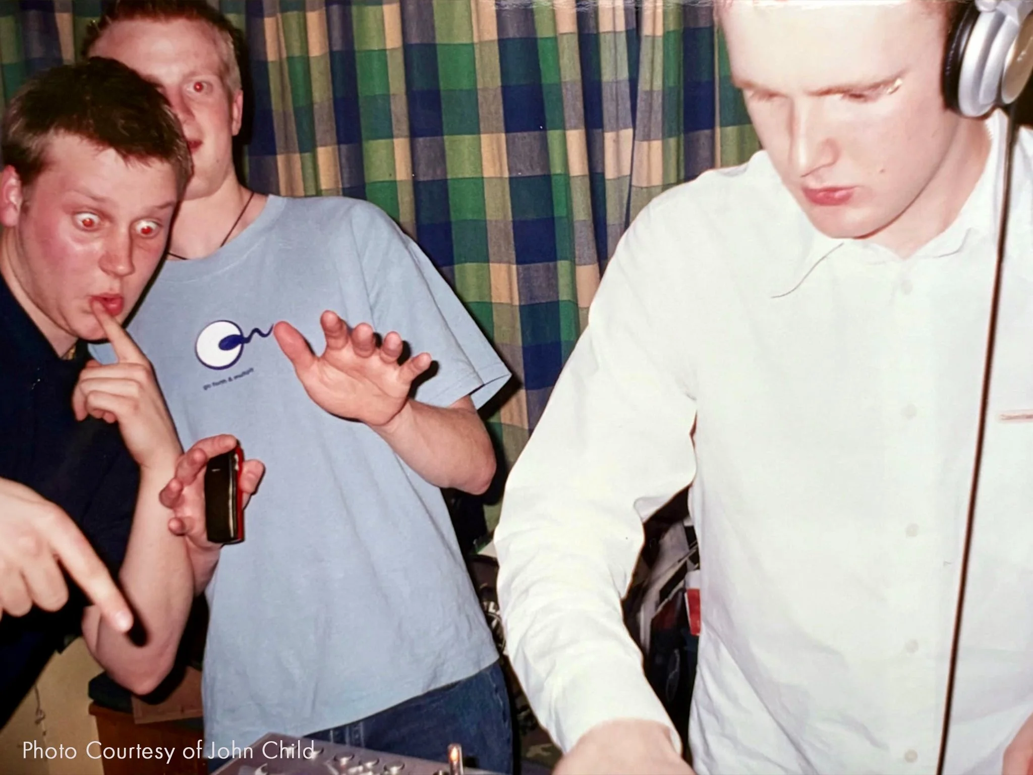 Three young men at a party or gathering, one is on the left, making a shushing gesture with his finger to his lips, another in the middle looks surprised with wide eyes and a hand raised, and the third on the right is focused on DJ equipment, wearing headphones and a white shirt.