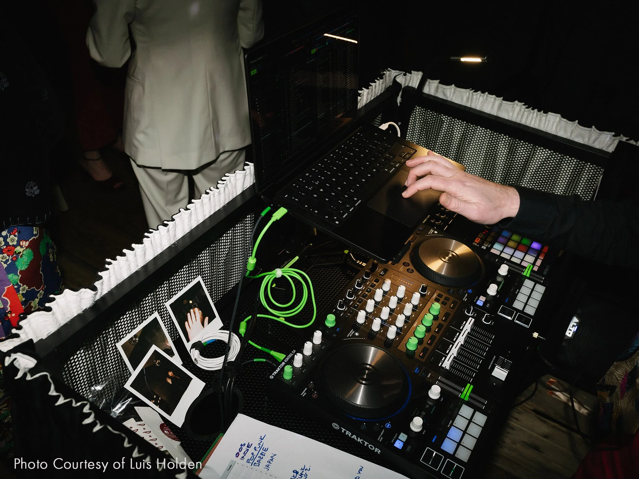 DJ mixing music at a DJ booth with turntables, a mixer, a laptop, and green headphones, in a dark environment with a person wearing a black shirt.