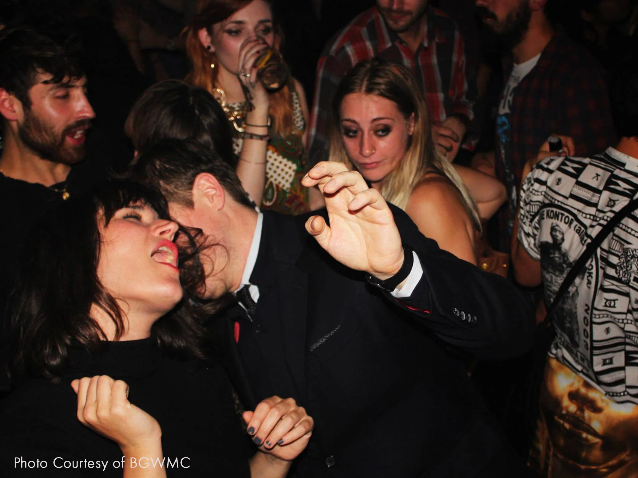 People dancing closely at a lively party, with women and men enjoying themselves and one woman drinking from a glass in the background.