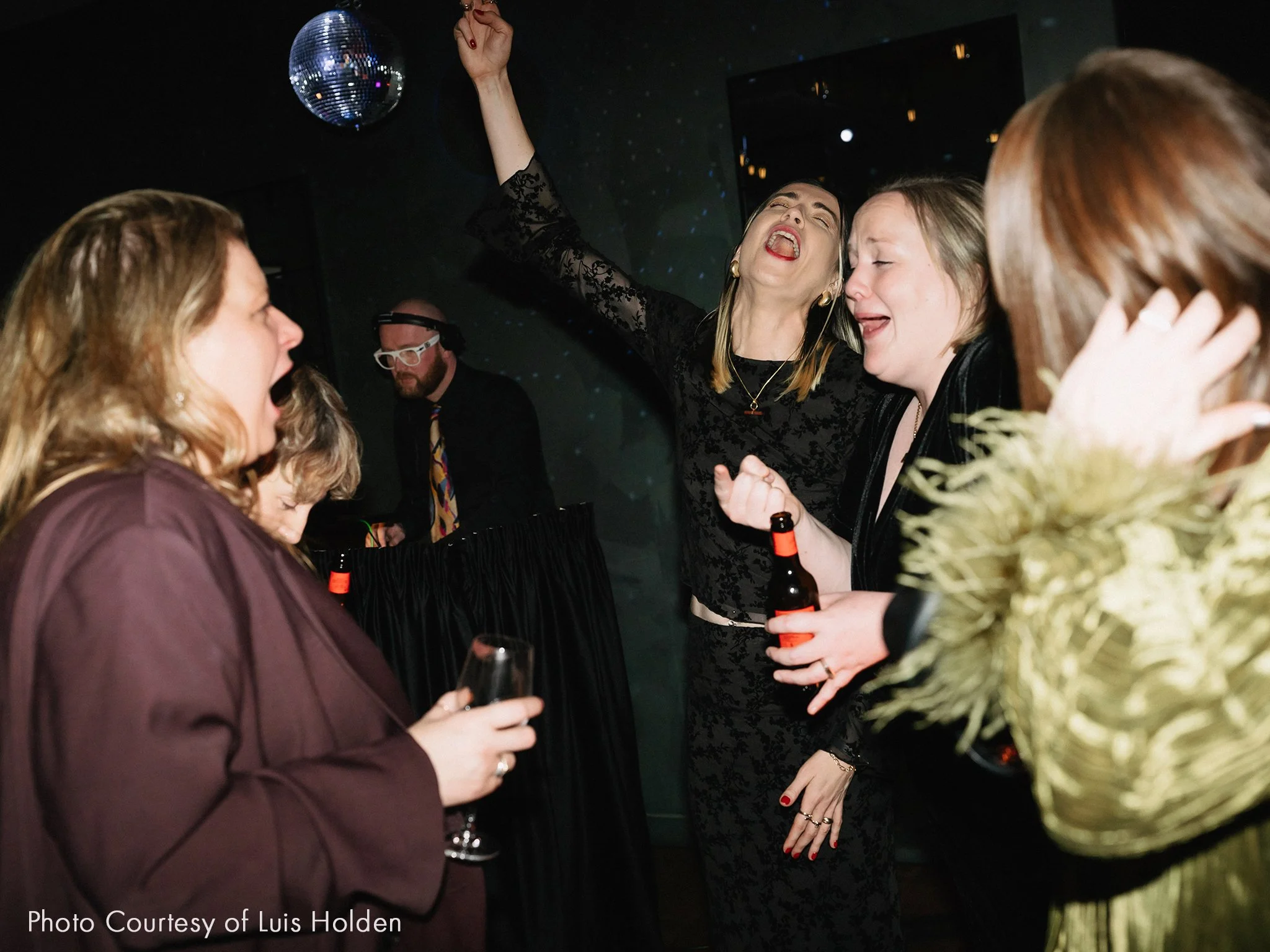 Group of women singing and dancing at a party with a disco ball, holding drinks, with a DJ in the background.