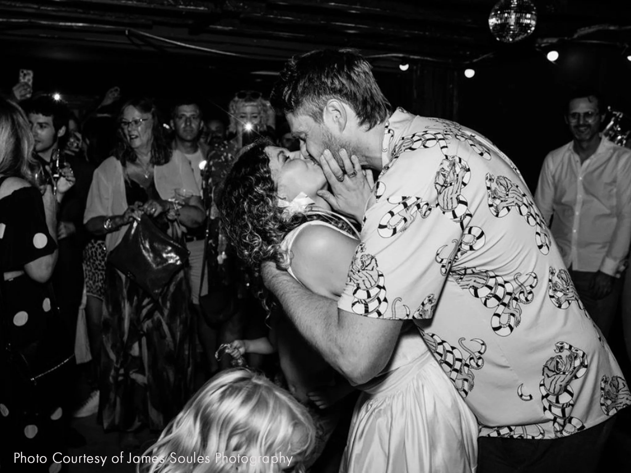 A newly married couple sharing a passionate kiss on a dance floor surrounded by friends and family.