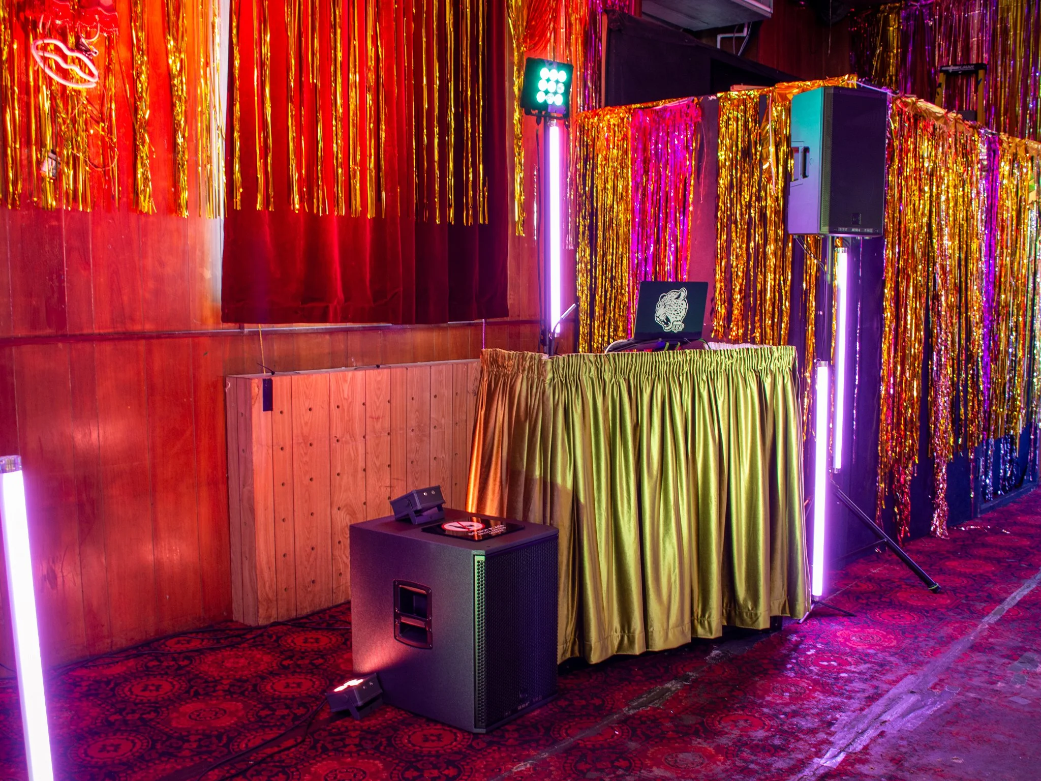 A empty indoor party scene with colorful decorations, shiny tinsel curtains, a DJ booth with a laptop showing a jaguar logo, large speakers, and neon lighting.
