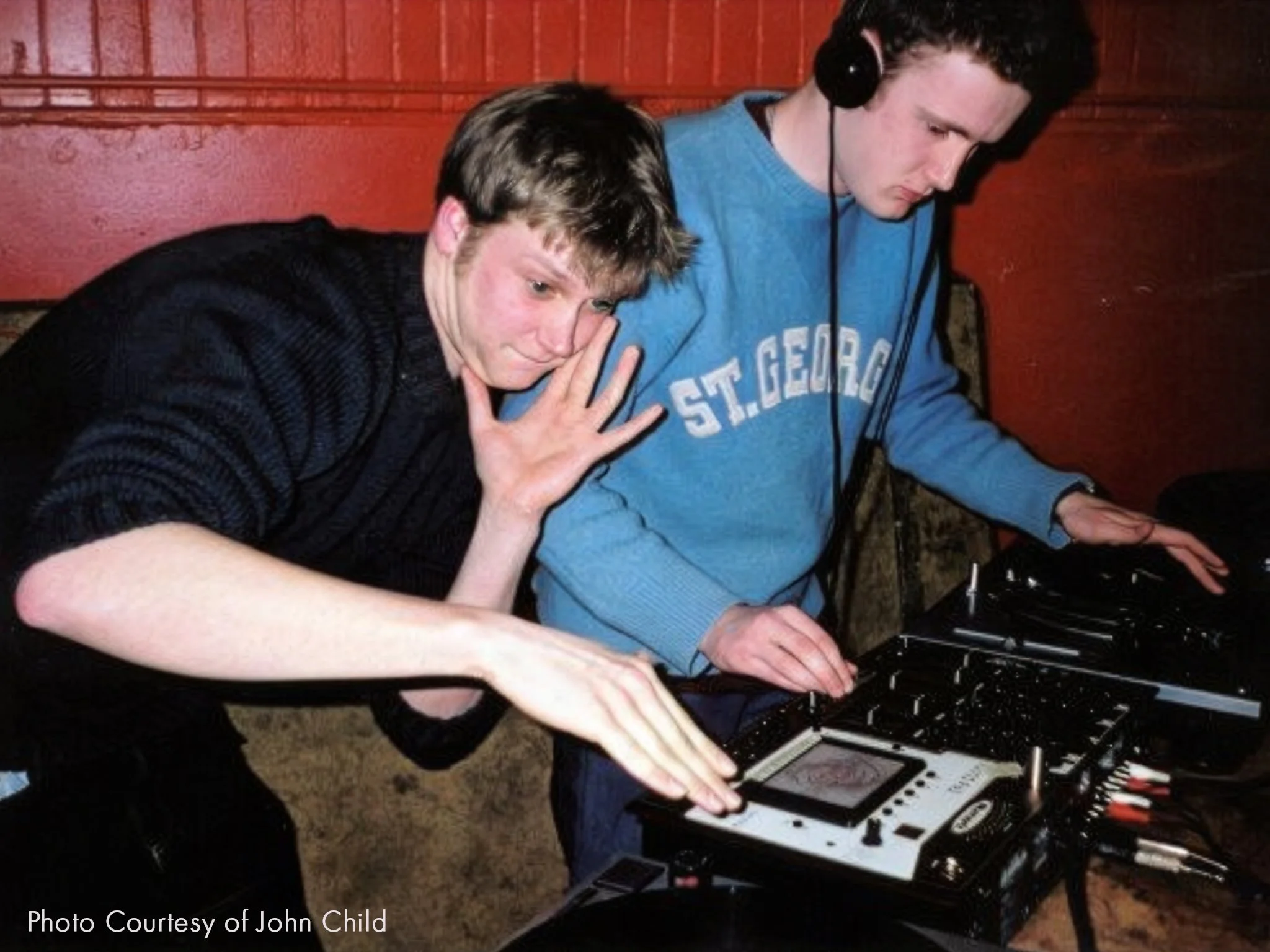 Two young men are DJing, with one wearing headphones and the other leaning over the turntable, both focused on the equipment.