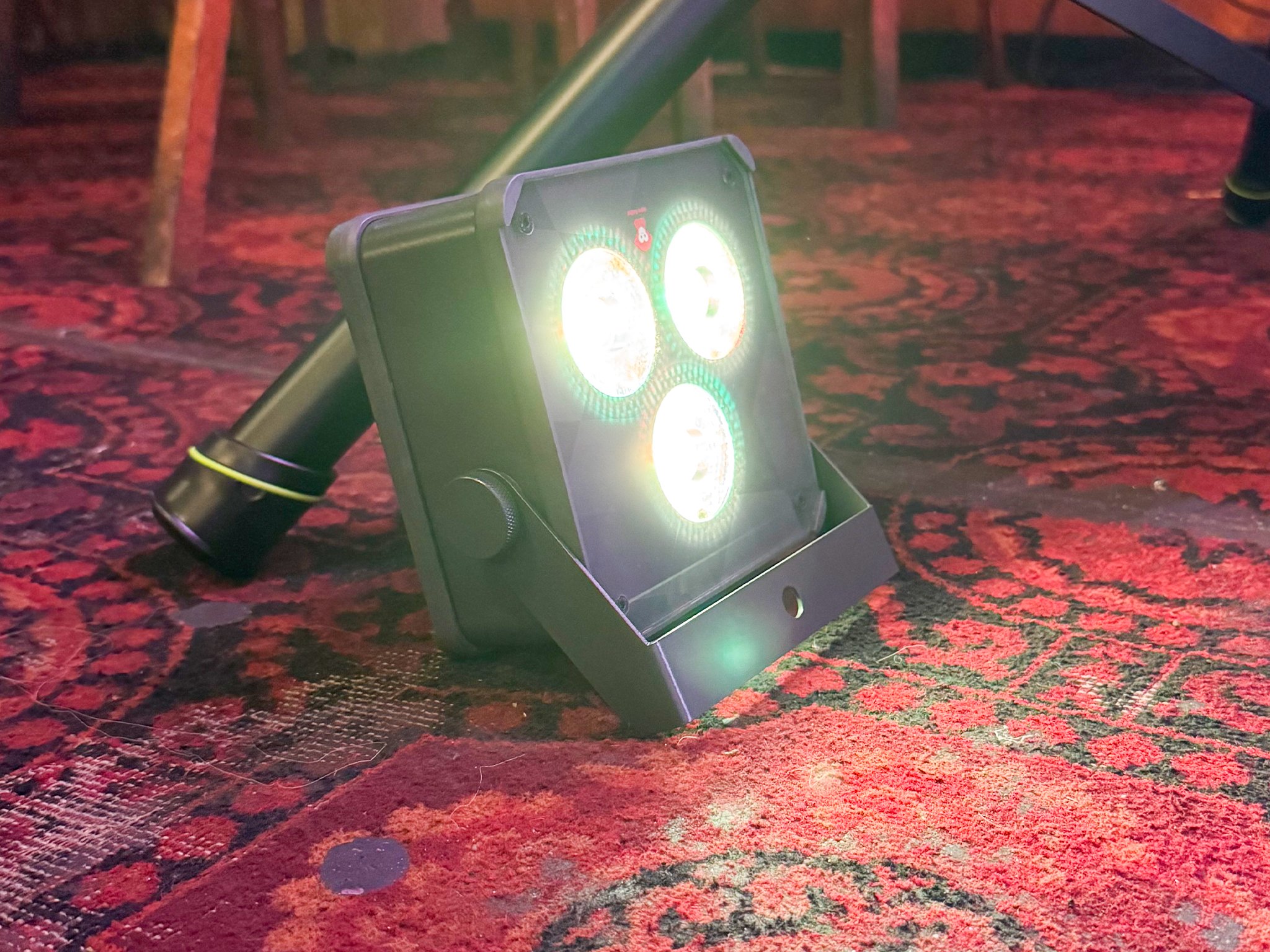 An Ape Labs Maxi V2 fixture with three bright, circular, multicolored lights, positioned on a red and black patterned carpet.