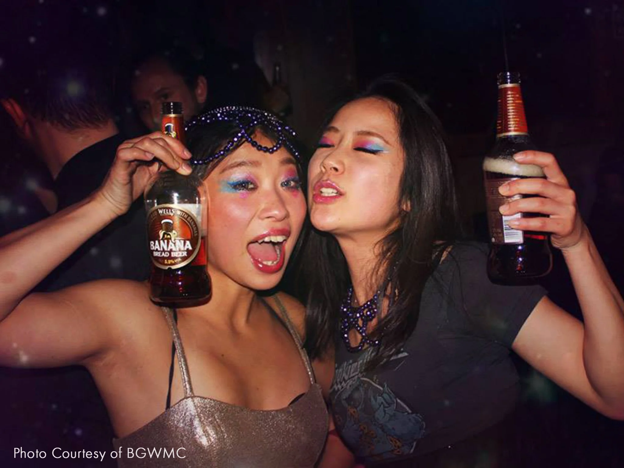 Two women with vibrant makeup and costumes holding beer bottles, celebrating at a party.