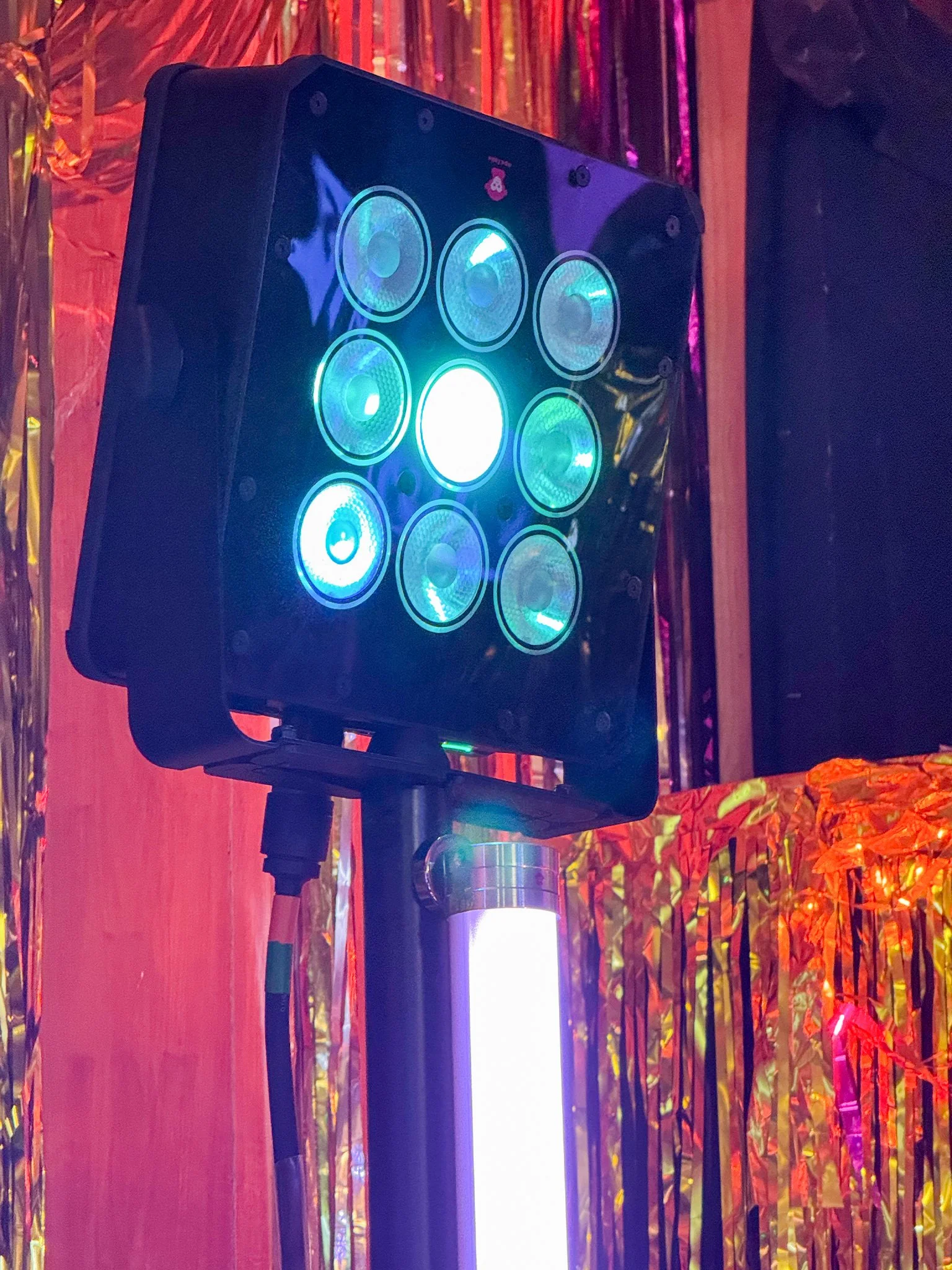 An Ape labs Marvel Spot fixture with multiple circular LED lights emitting blue and white light, mounted on an adjustable stand with a glowing Neon tube light, set against colorful foil decorations.