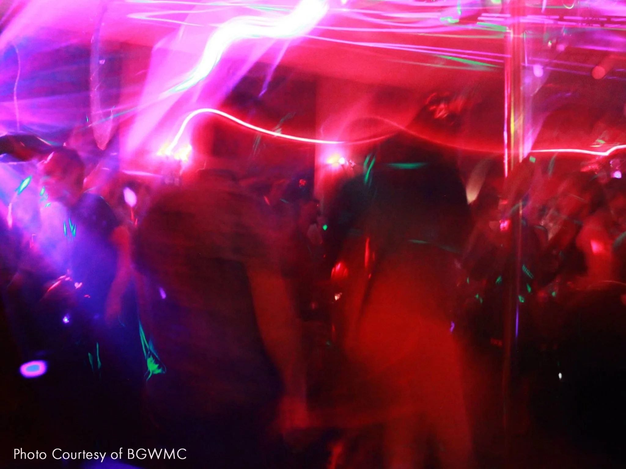 Blurred photo of a dance party or nightclub scene with colorful neon lights and multiple people dancing.