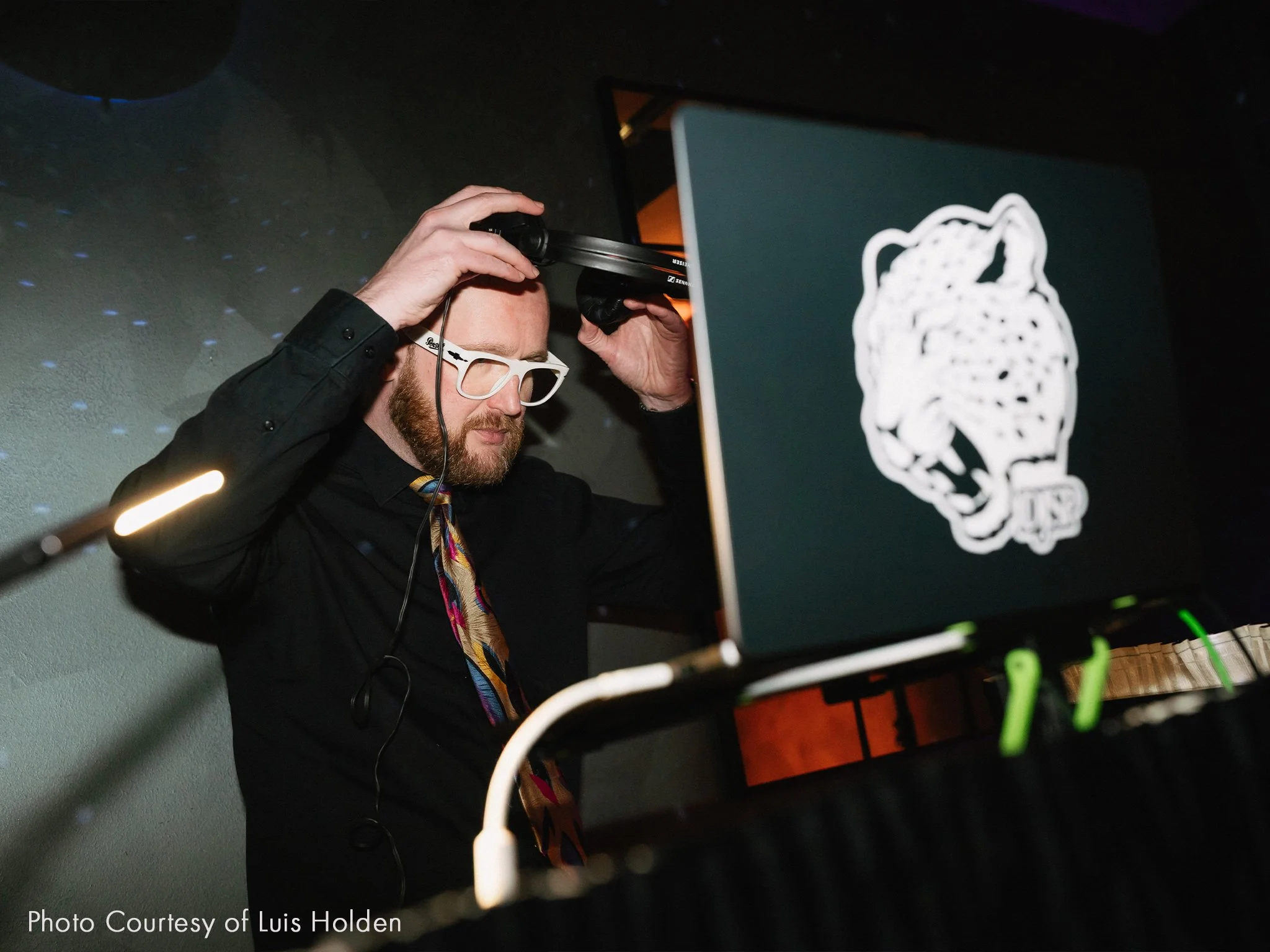 A DJ with white glasses and a black shirt adjusting headphones behind a turntable, with a laptop displaying a jaguar head logo.