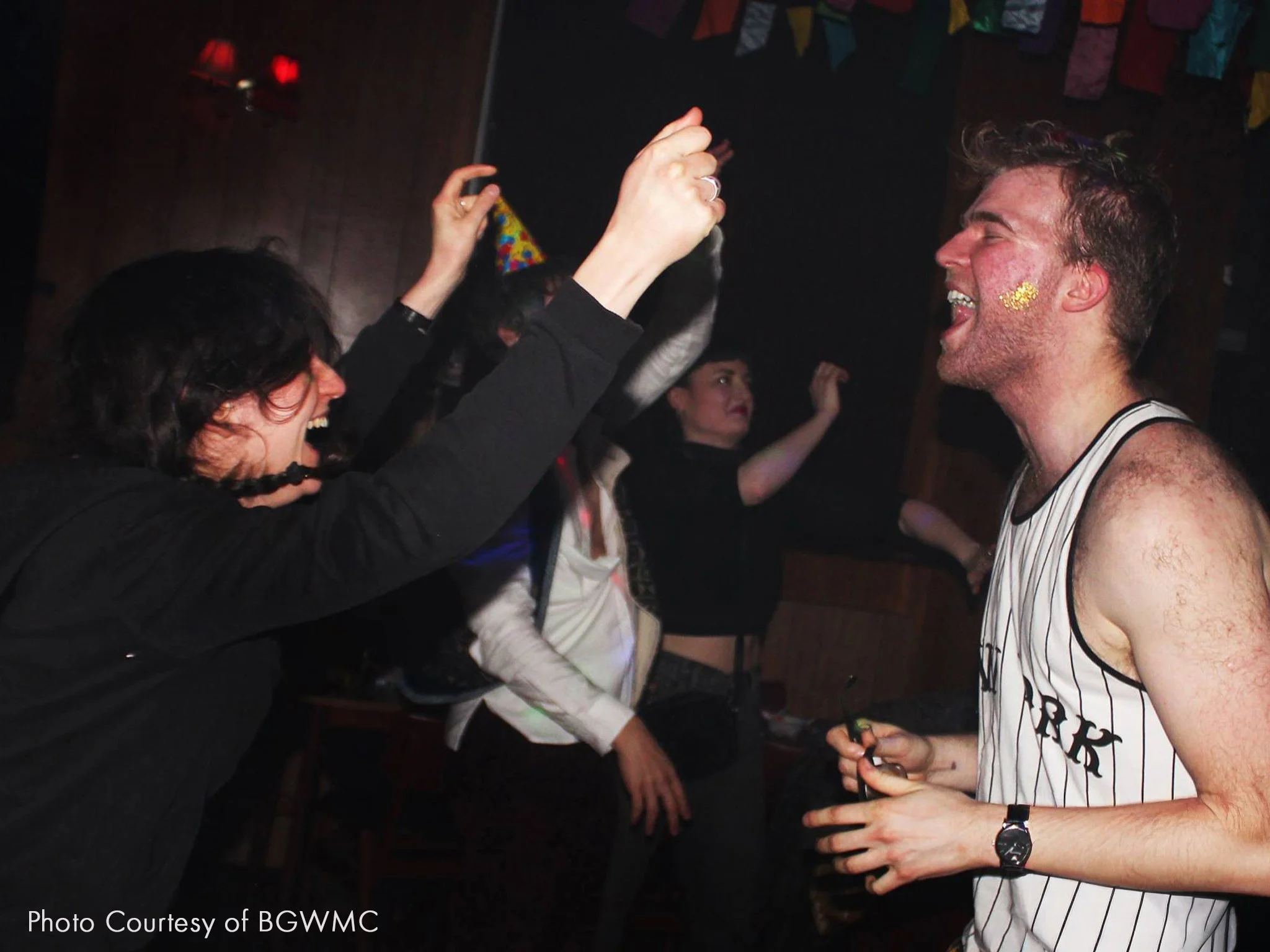 People celebrating and dancing at a party, with one person wearing a striped shirt and another with a party hat, in a dimly lit room decorated with colorful banners.