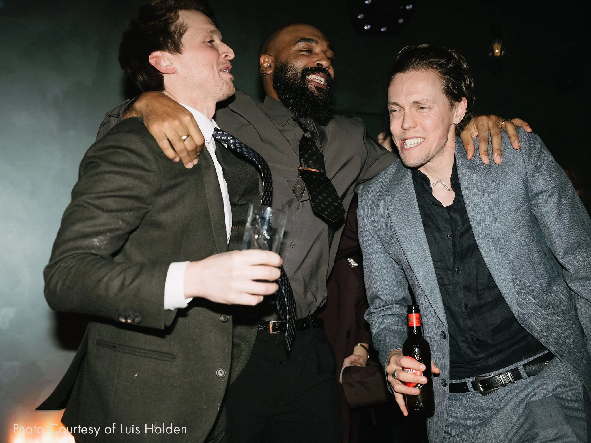 Three men in suits enjoying drinks and laughing at an indoor party.
