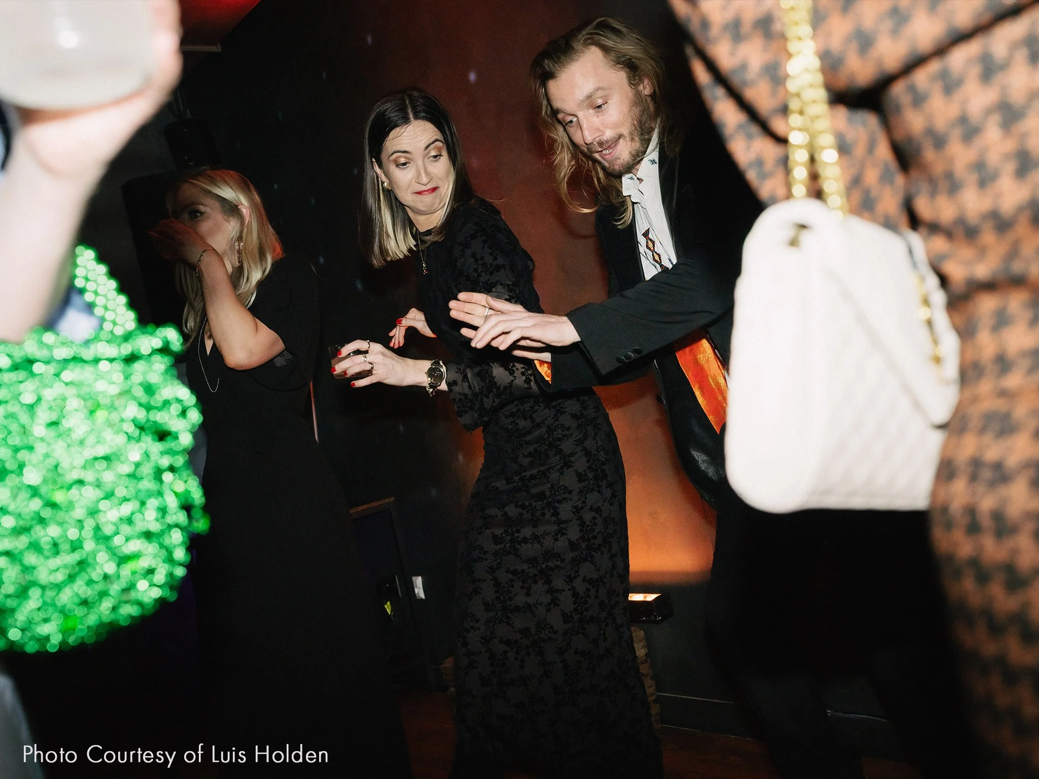People dancing at a party or celebration with one woman in a black lace dress and a man in a black jacket, while others are partially visible around them.