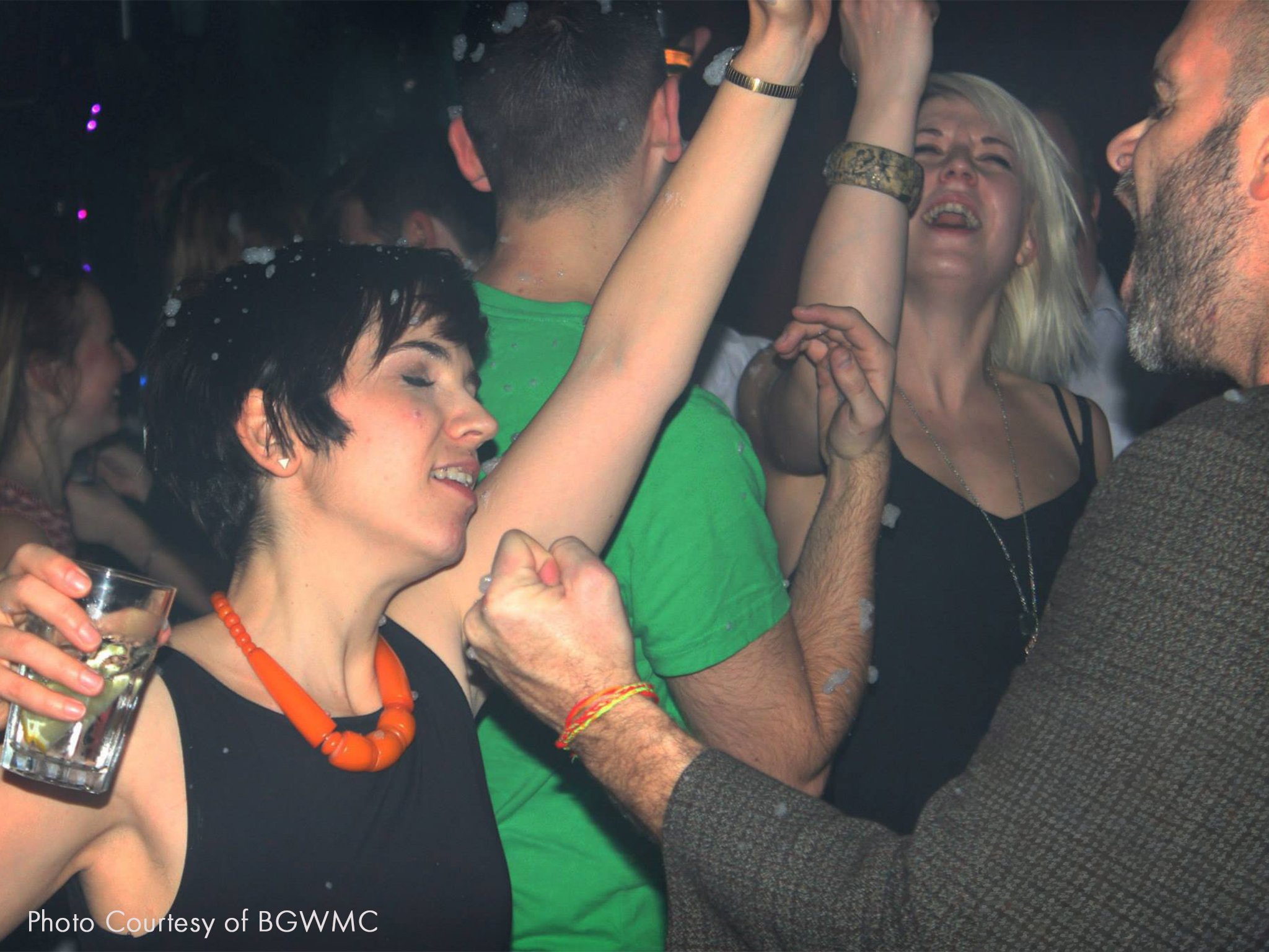 People dancing and enjoying themselves at a party, with close-up focus on a woman with short black hair and orange jewelry, a woman with blonde hair, and a man with a beard, are all having fun.