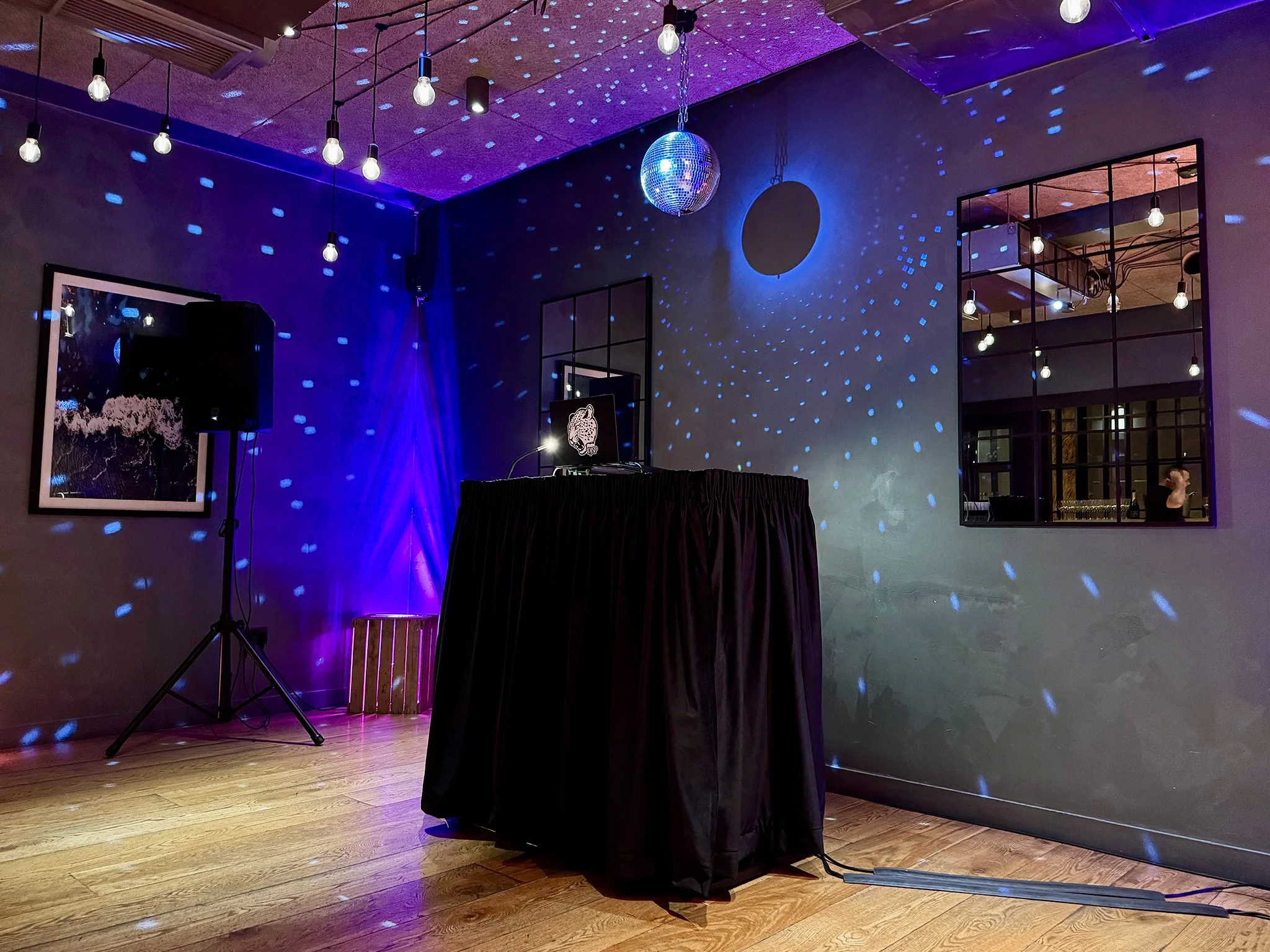 A dimly lit dance or event space with disco ball, blue light projections on walls and ceiling, a DJ setup on a black table, and string lights hanging from the ceiling.