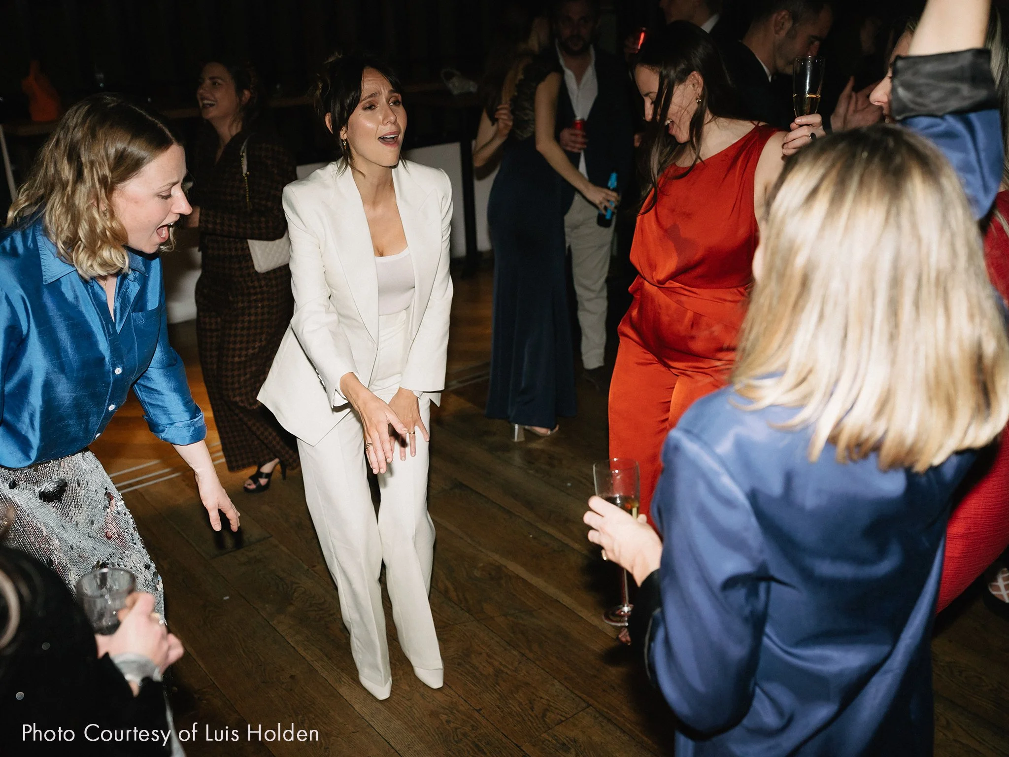 Group of people dancing and singing at a wedding celebration, with women holding glasses of champagne.