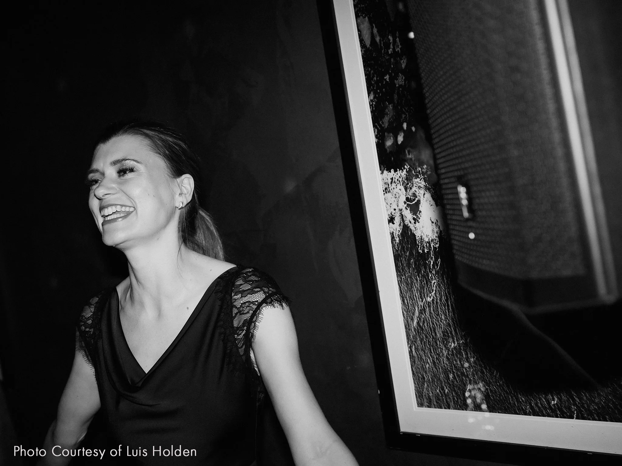 A woman smiling and laughing, dressed in a black top with lace sleeves, standing next to a loud speaker. The image is in black and white.