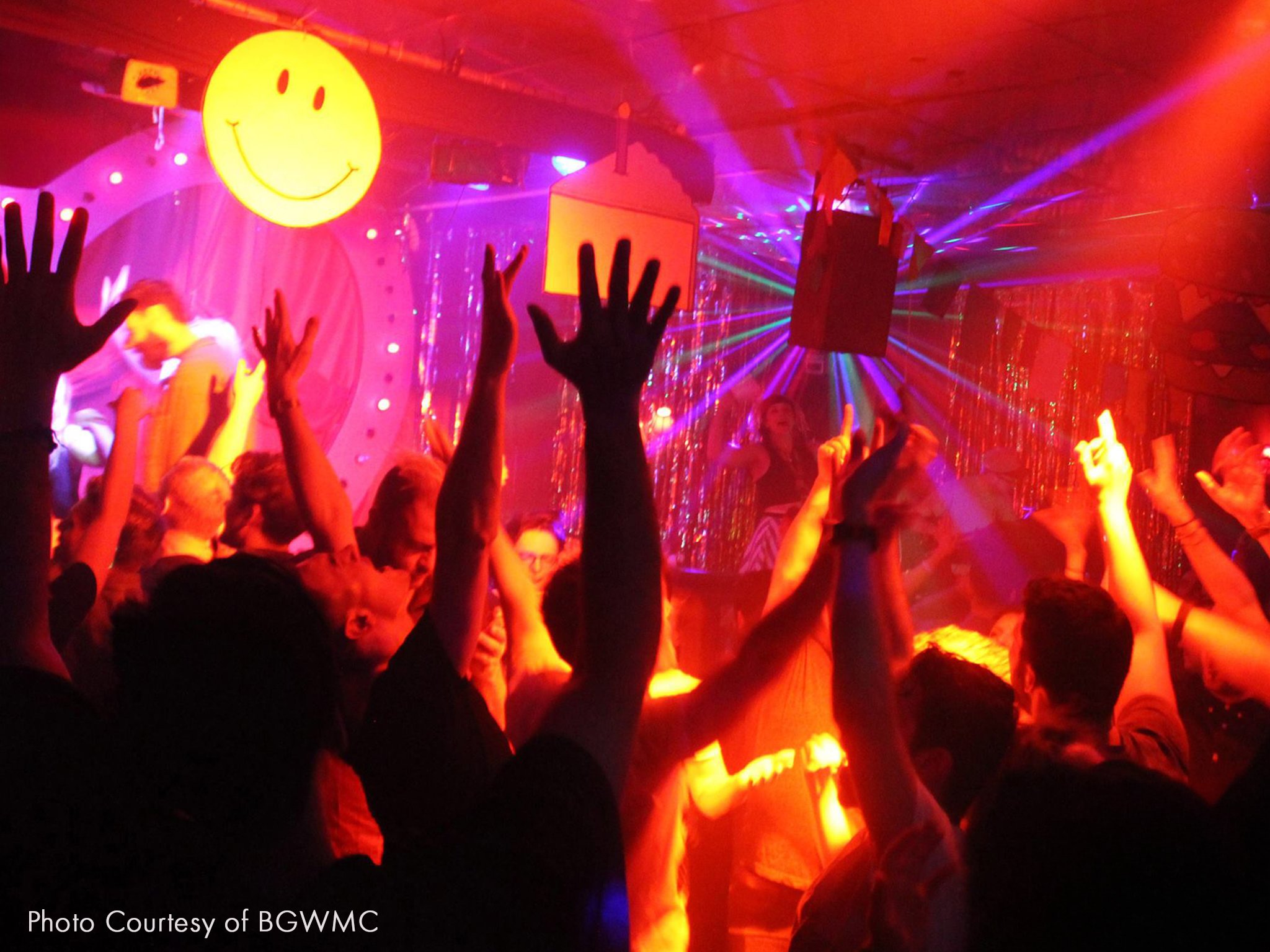 People dancing at a lively party with colorful lights, a large yellow smiley face, and festive decorations. They have their hands up in the air as their favourite track plays.