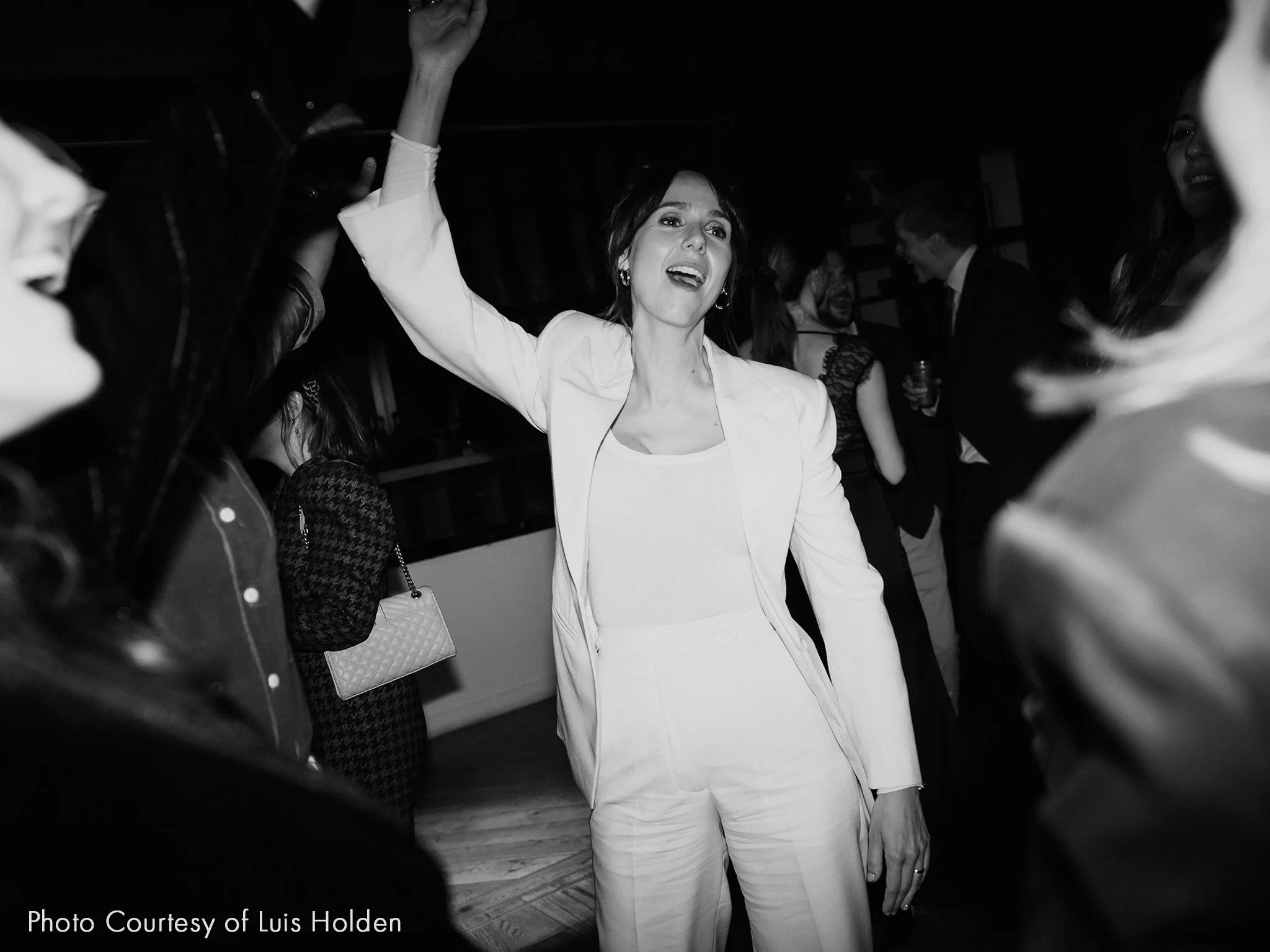 A bride in a white trouser suit dancing at her wedding with other guests in the background.