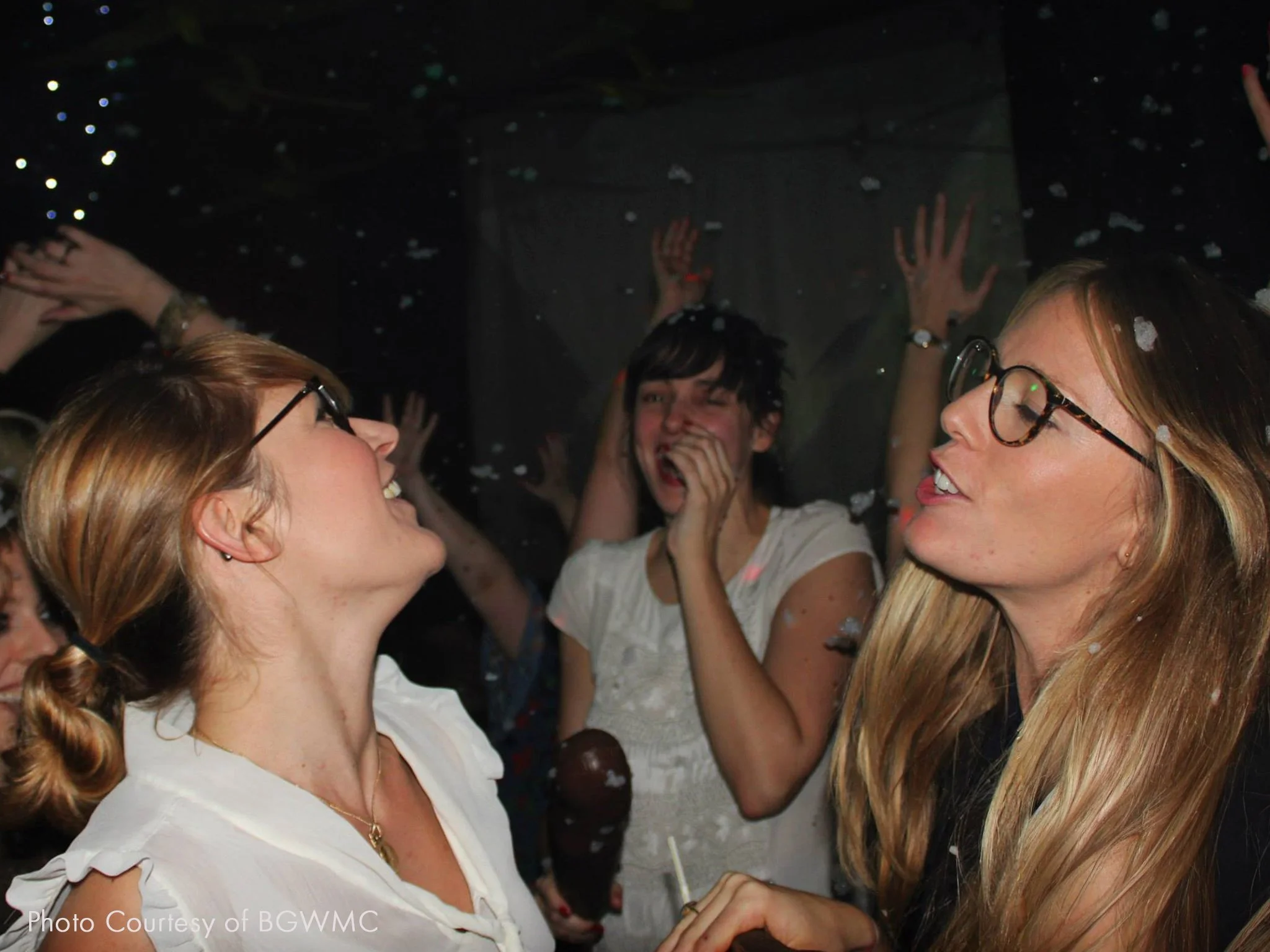 People celebrating at a party, with some smiling and others talking, in a lively indoor setting with confetti in the air.