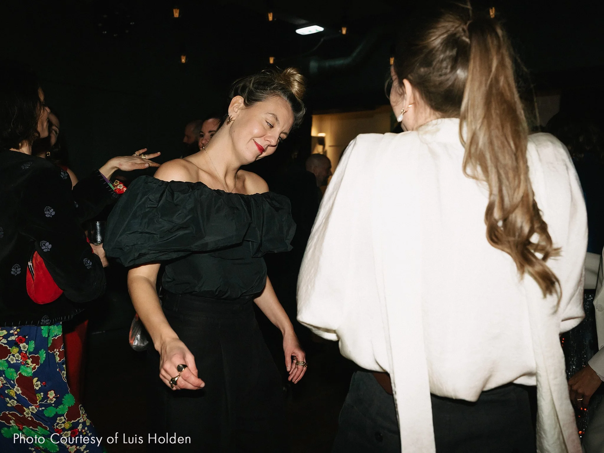 Two women dancing and enjoying themselves at a social event in a dimly lit room. The woman on the left has light skin, her hair in a bun, wearing a black off-shoulder top with puffy sleeves, and rings on her fingers. The woman on the right has light skin, long wavy hair, and is wearing a white top with puffed sleeves and black pants.