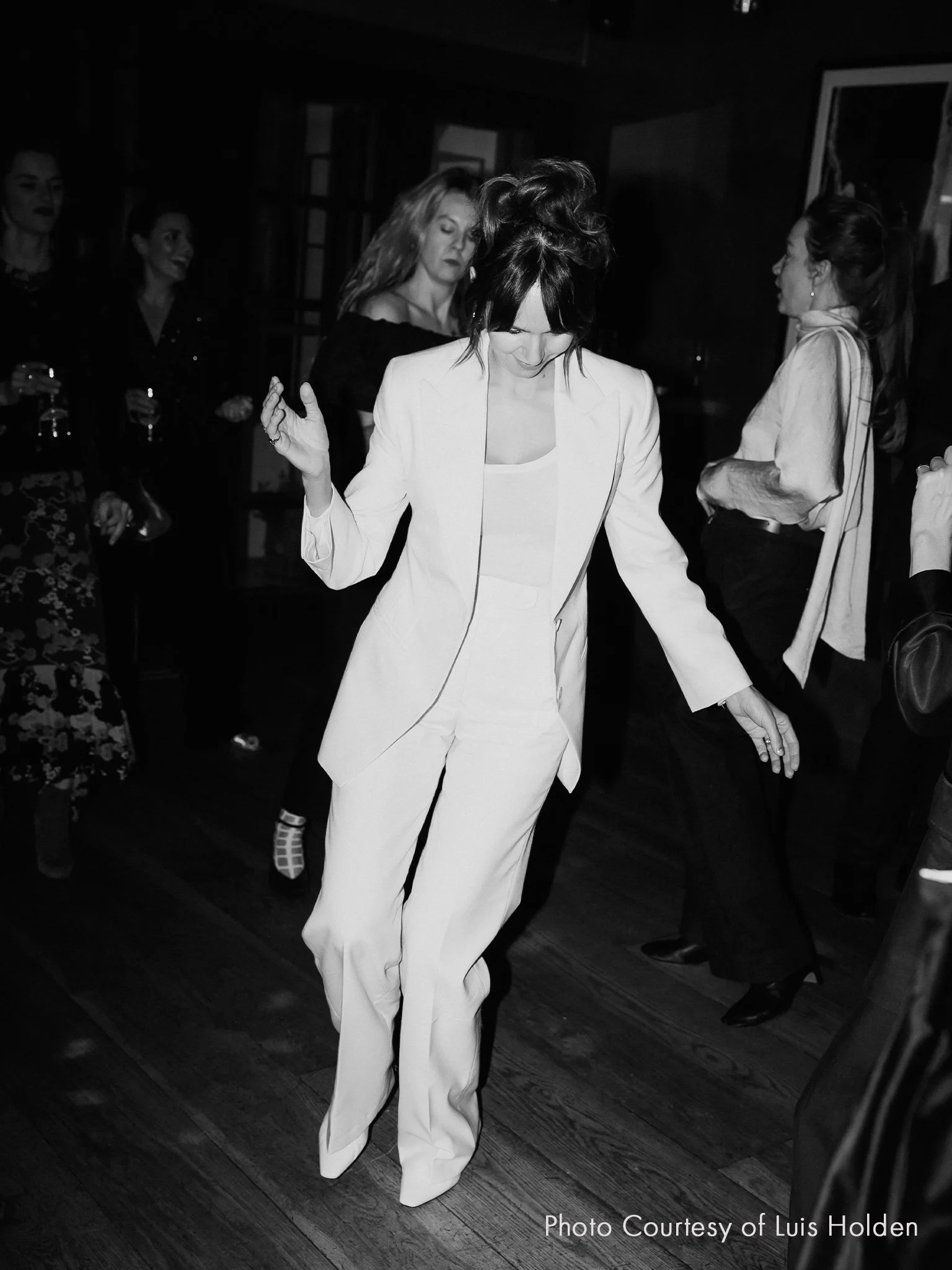 A bride dancing in a white trouser suit at her wedding, with others in the background enjoying the event.