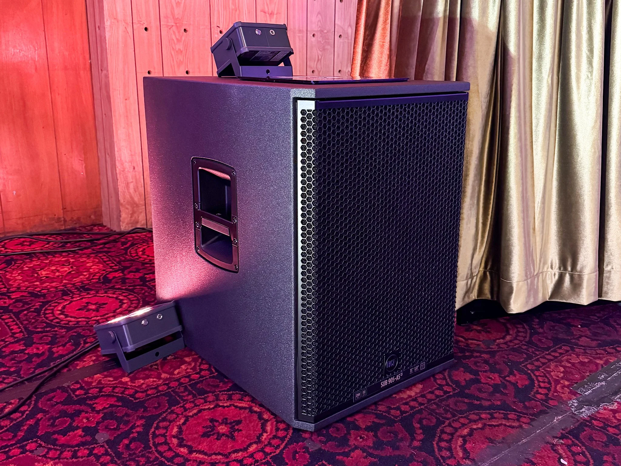 Large black speaker on a red patterned carpet with a wooden wall and golden curtains in the background.