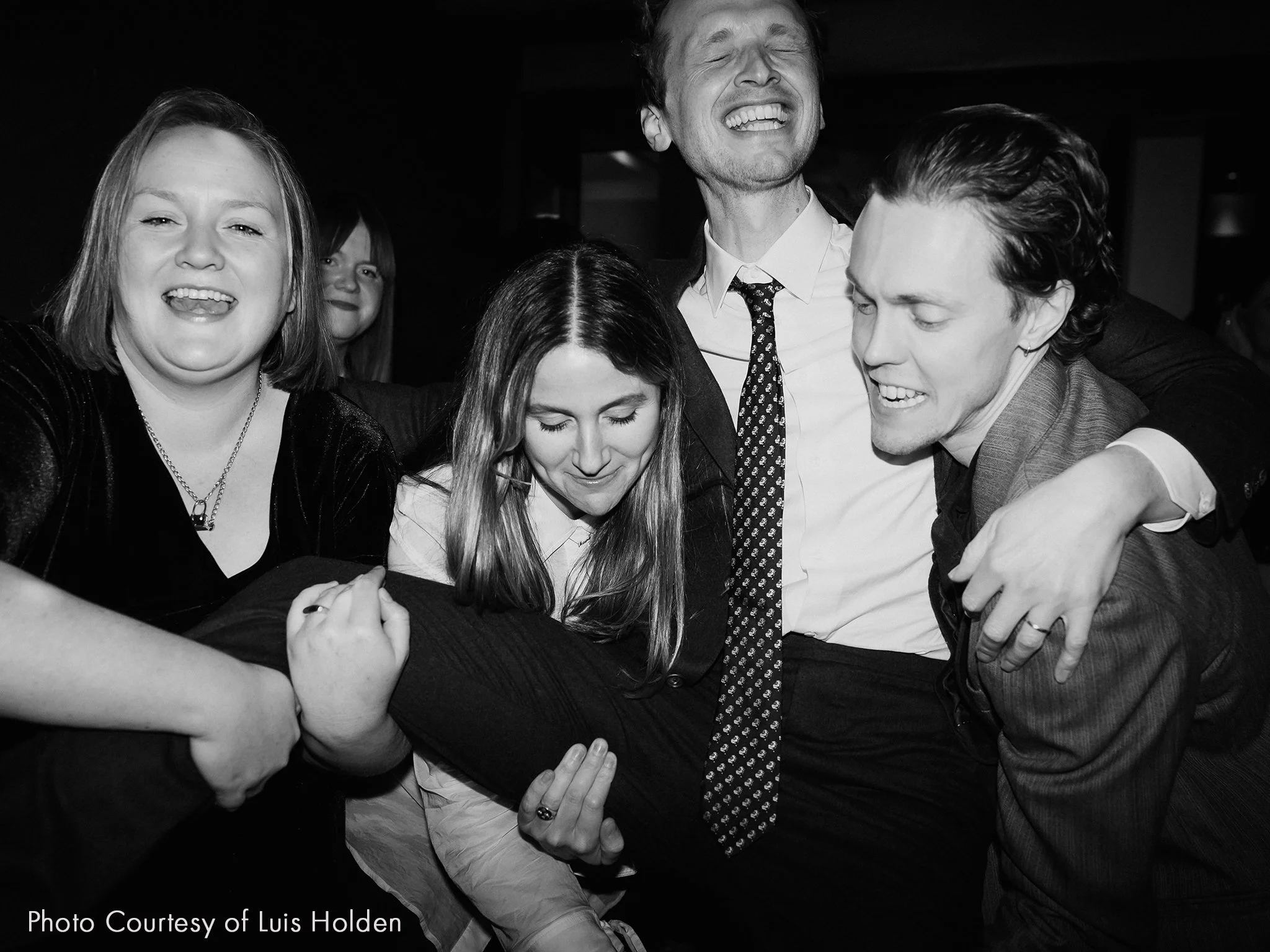 Group of five people celebrating and laughing closely together, with one person being held horizontally in a group hug. Black and white photo.