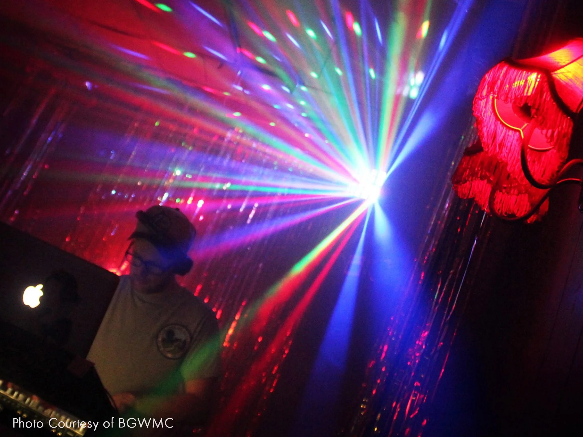 Person with glasses and baseball cap DJing in front of colorful laser lights. A disco light is causing a colourful array.