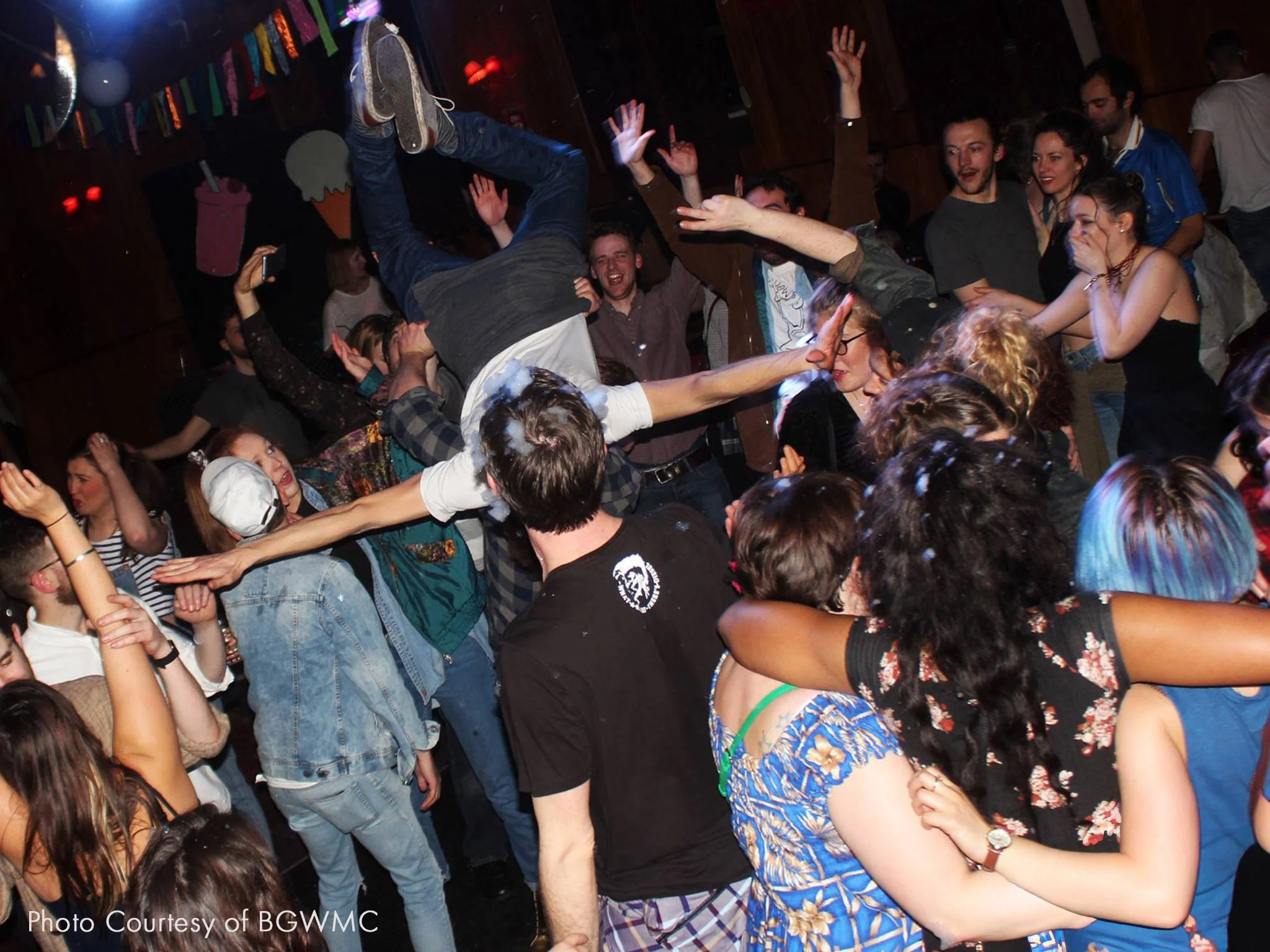 People dancing and celebrating at a crowded party, with a man being lifted into the air by a group of friends.