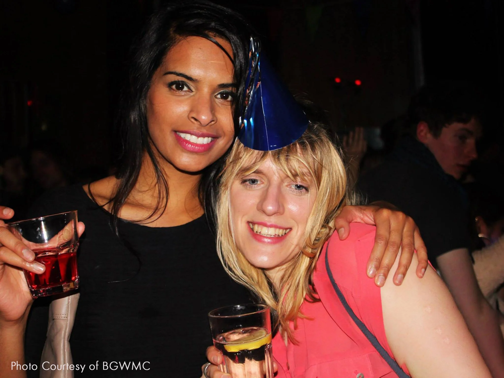 Two women at a celebration, one wearing a blue party hat, holding glasses of pink and yellow drinks, smiling, with a dark background and other people in the distance.