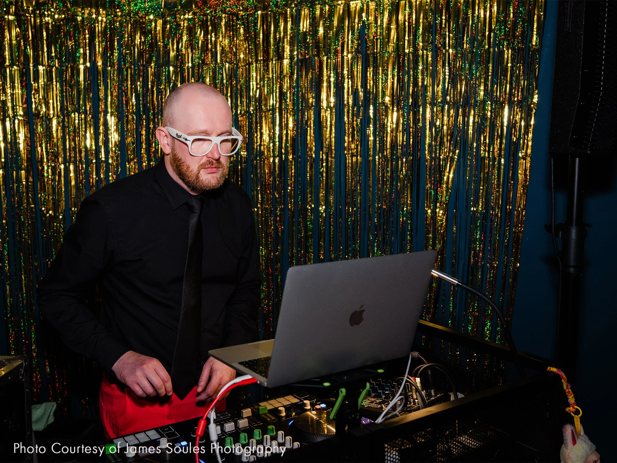 A DJ wearing white glasses, black shirt, and red pants is performing at a party with a gold tinsel backdrop.