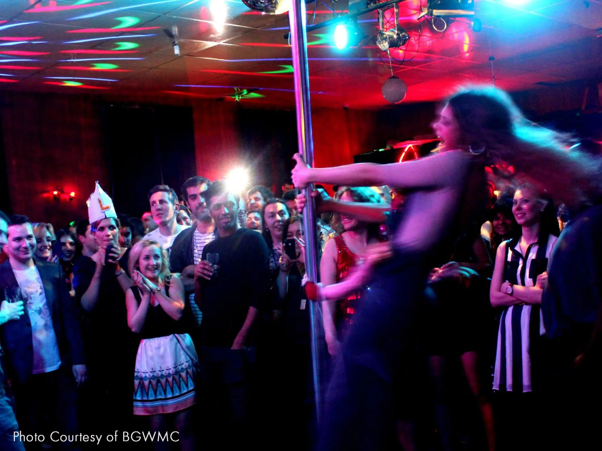 A woman dancing on a pole at a crowded party, with guests watching and smiling, under colorful lighting.